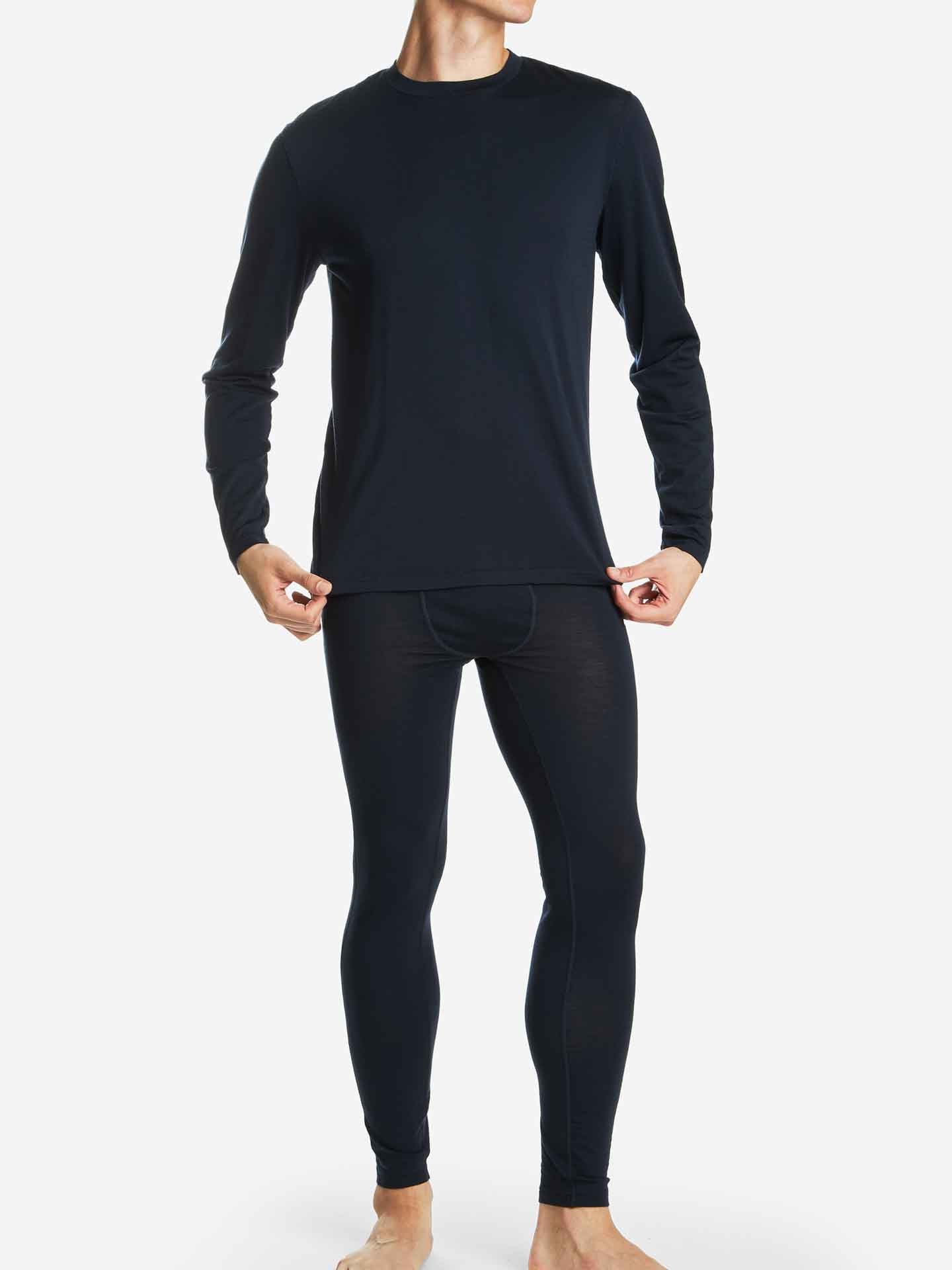 A person wearing the Snø Merino Long Sleeve Men from We Norwegians, along with matching pants, stands on a light background, pulling the bottom of the shirt slightly downward. The outfit appears to be a stretchy fabric base-layer set, ideal for activewear or thermal comfort.