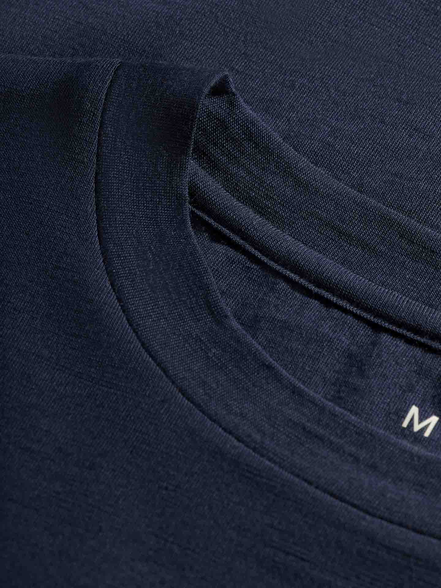 A close-up of the Snø Merino Long Sleeve Men's dark blue collar highlights layered, stretchy fabric and intricate stitching. The partially visible "M" on the inside label indicates its comfort as a base layer, likely made from fine Merino wool by We Norwegians.