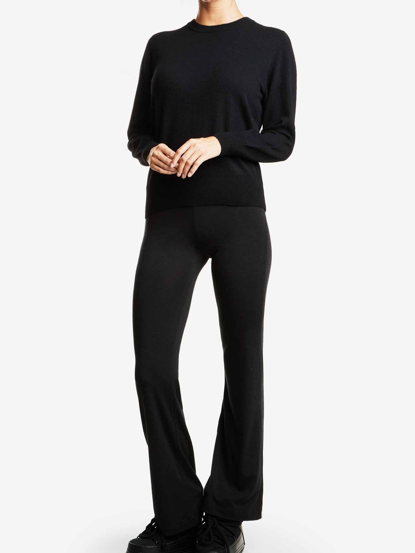 A person wearing a We Norwegians Blefjell Cashmere Crewneck Women in black and black flared pants stands against a plain white background. The outfit, crafted from pure cashmere, exudes casual comfort. Their hands are clasped together in front of them, showcasing this luxury staple.