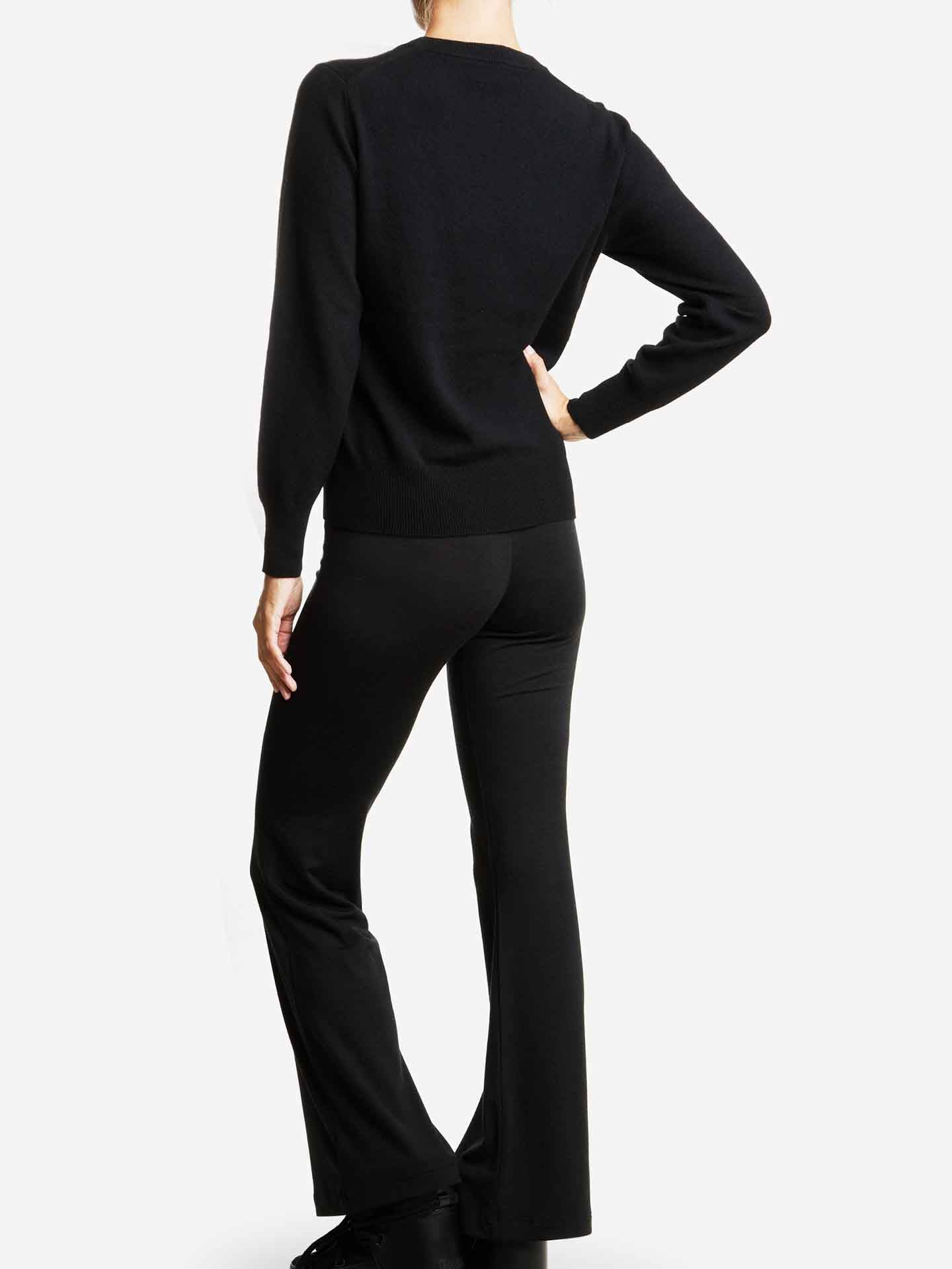 A person wearing a black We Norwegians Blefjell Cashmere Crewneck Women and black flared pants stands with their back to the camera. Their left hand rests on their hip, and they appear to be in a neutral pose against a plain, light-colored background.