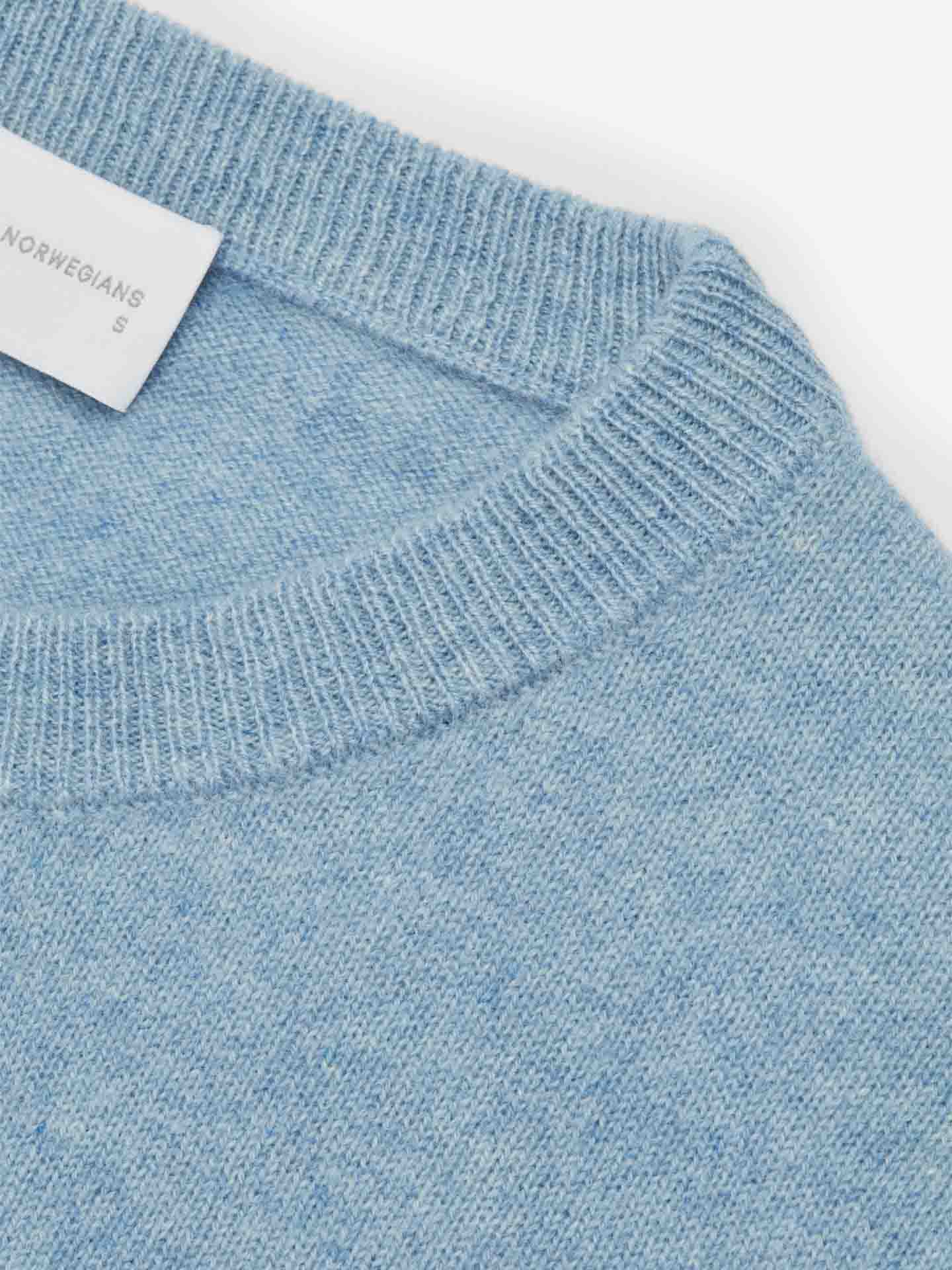 Close-up of the Blefjell Cashmere Crewneck Women by We Norwegians, featuring a light blue knit with a ribbed, round neckline. A white clothing tag with partially visible text is attached to the inside of the neckline, indicating the size "S." This luxury staple, crafted from pure cashmere, embodies elegance and comfort.