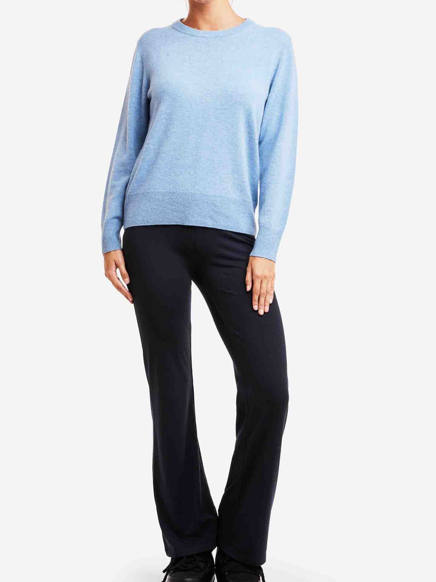 A person standing against a white background, wearing a light blue We Norwegians' Blefjell Cashmere Crewneck Women and black pants. The individual is posing with one hand resting on their hip and the other arm relaxed by their side. The face is not visible in the image, letting the luxury staple speak for itself.