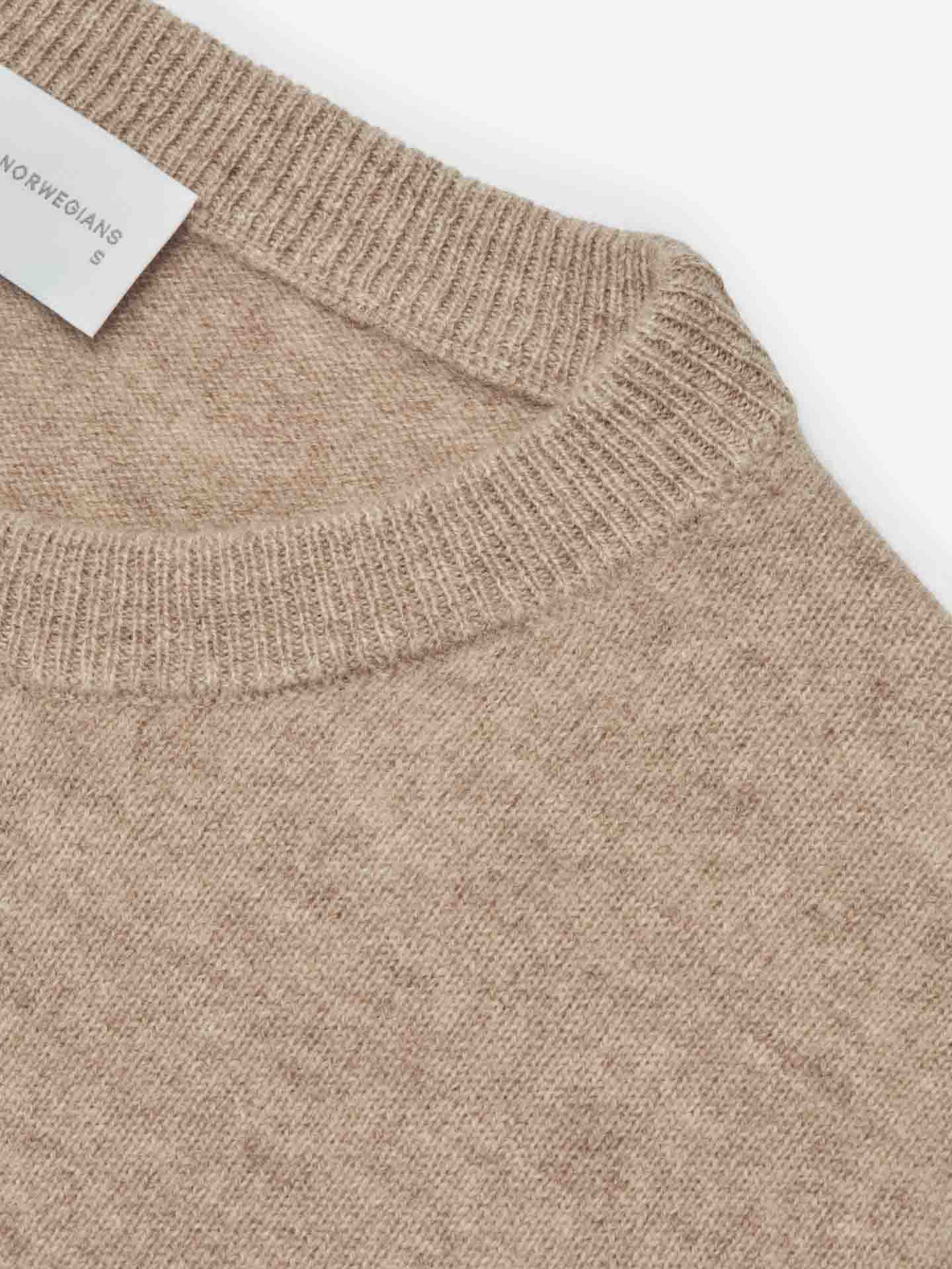 Close-up of a beige We Norwegians Blefjell Cashmere Crewneck Women sweater showing the neckline and a small portion of the shoulder seam. The pure cashmere knit fabric appears soft and cozy. A partially visible white clothing tag is attached near the neckline, emphasizing this luxury staple.