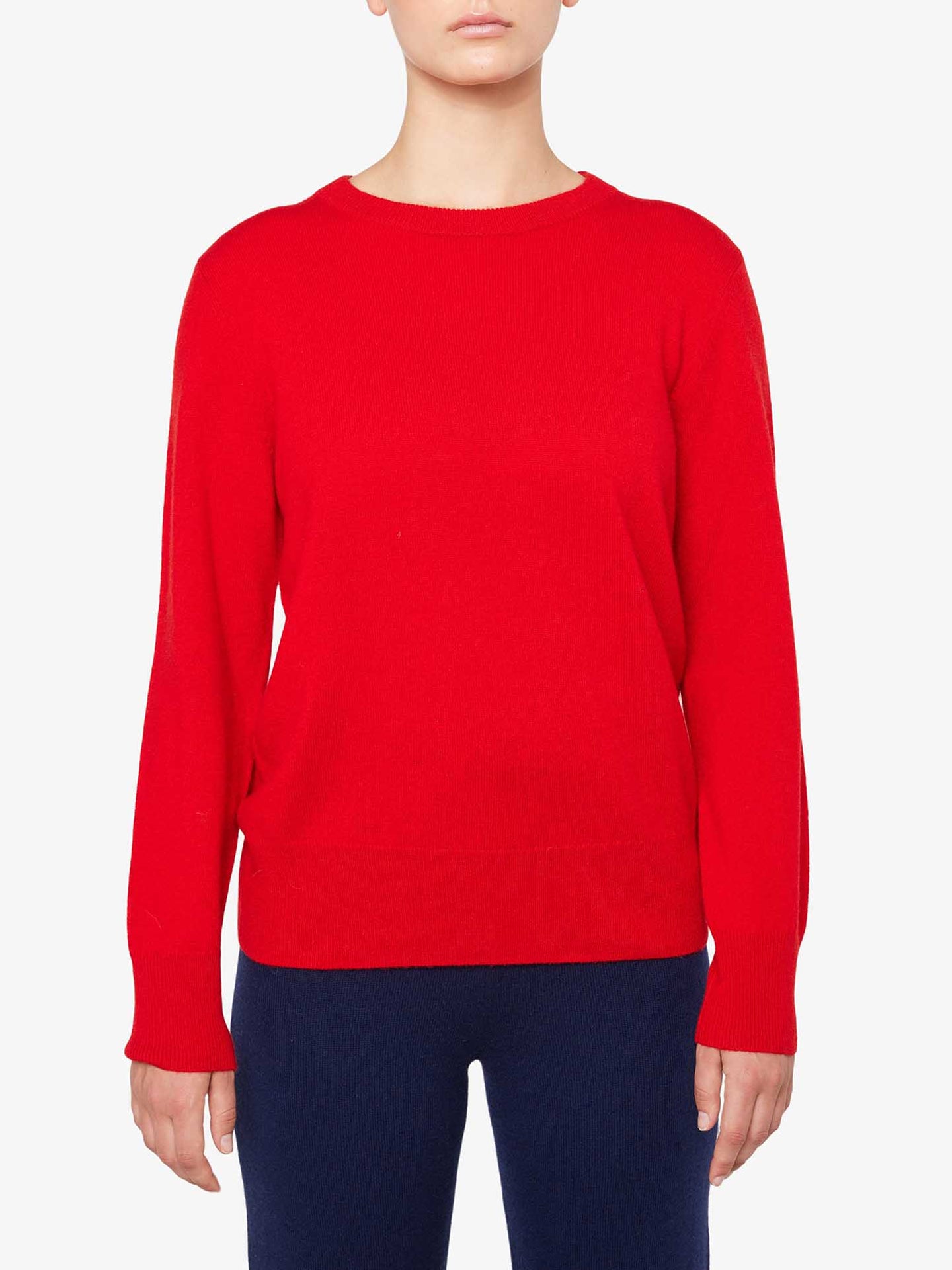 A person wearing a bright red Blefjell Cashmere Crewneck Women sweater from We Norwegians and dark blue pants stands against a plain white background. The luxury staple features a round neckline and a relaxed fit. The person's face is not visible.