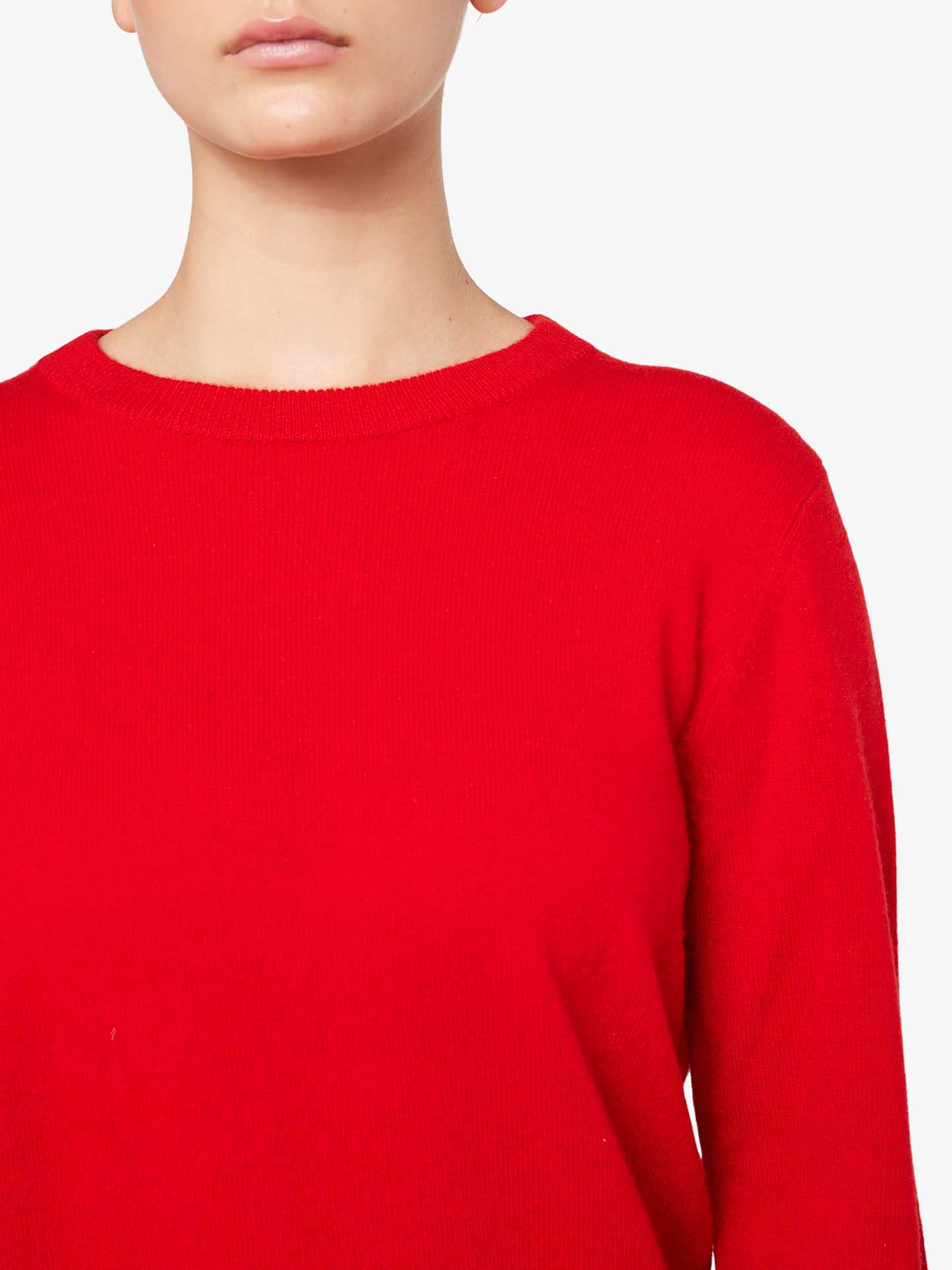 A person is wearing a bright red Blefjell Cashmere Crewneck Women sweater from We Norwegians. The image is cropped at the nose level, not showing the person's full face. The background is plain white, making this luxury staple in pure cashmere stand out prominently.