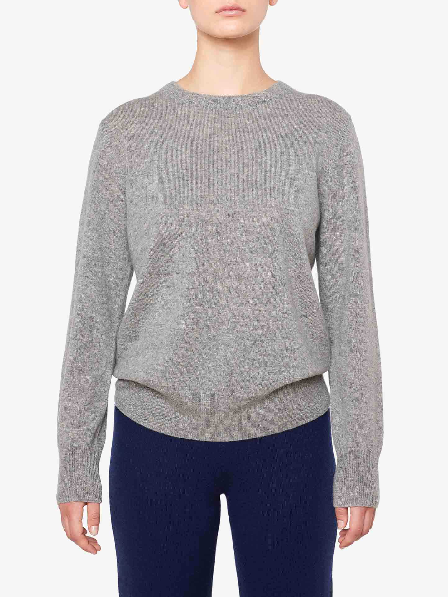 A person is standing against a plain white background, wearing a light gray We Norwegians Blefjell Cashmere Crewneck Women with long sleeves and dark blue pants. The person’s arms are relaxed at their sides. The image is cropped just below the shoulders and mid-thigh, showcasing this luxury staple in pure cashmere.