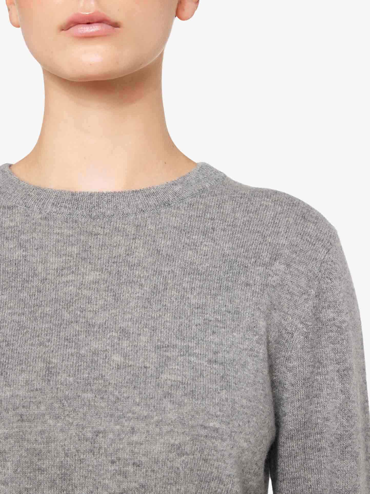 A close-up image of a person wearing a soft, gray We Norwegians Blefjell Cashmere Crewneck Women sweater. The photograph is cropped at the nose, showing only the lower part of the face, neck, and upper torso against a white background. The texture of this luxury staple knit fabric is clearly visible.