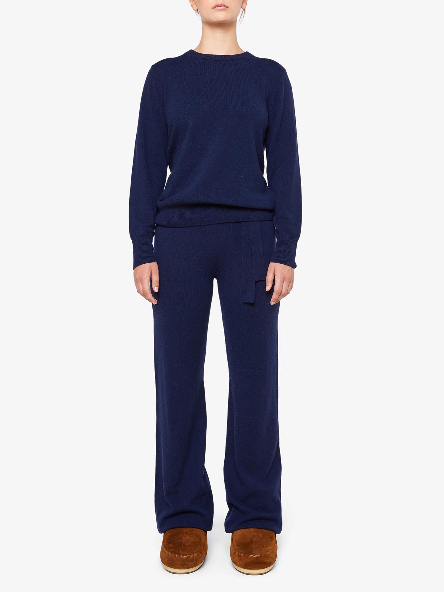 A person is wearing a dark blue Blefjell Cashmere Crewneck Women by We Norwegians, paired with matching dark blue wide-leg pants featuring a tied waist. They are also wearing brown slip-on shoes, set against a plain white background. This ensemble, crafted from pure cashmere, exudes timeless luxury and comfort.