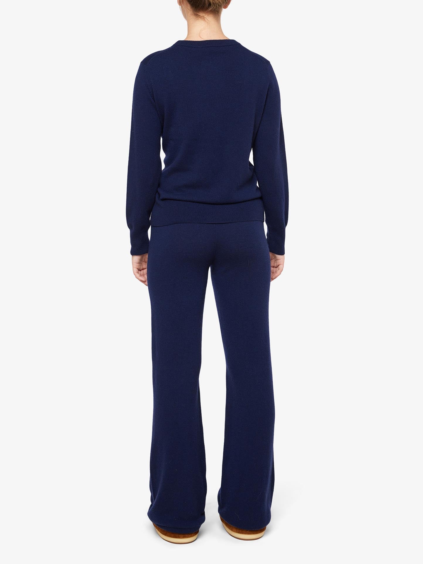 A person is standing with their back to the camera, dressed in a coordinated navy blue set from We Norwegians, featuring the Blefjell Cashmere Crewneck Women and matching wide-legged pants. The luxurious ensemble is complemented by light brown shoes, set against a plain white background.