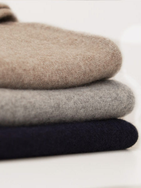 A stack of three folded Blefjell Cashmere Crewneck Men sweaters from We Norwegians in neutral tones. The top sweater is beige, the middle one is gray, and the bottom one is navy blue. Made of pure cashmere yarn, they appear soft and cozy, neatly layered on a white surface—a versatile luxury staple for any wardrobe.