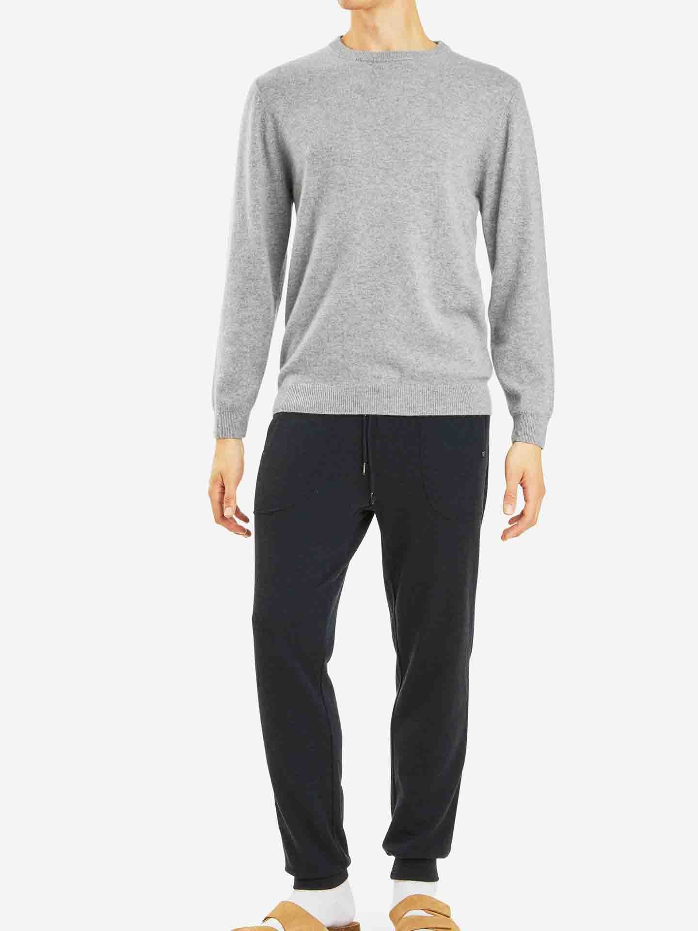 A person is standing against a plain white background, wearing a light gray Blefjell Cashmere Crewneck Men sweater by We Norwegians, dark navy sweatpants, white socks, and brown slippers. This versatile luxury staple showcases pure cashmere yarn for an elegant look. The person's face is not visible in the image.