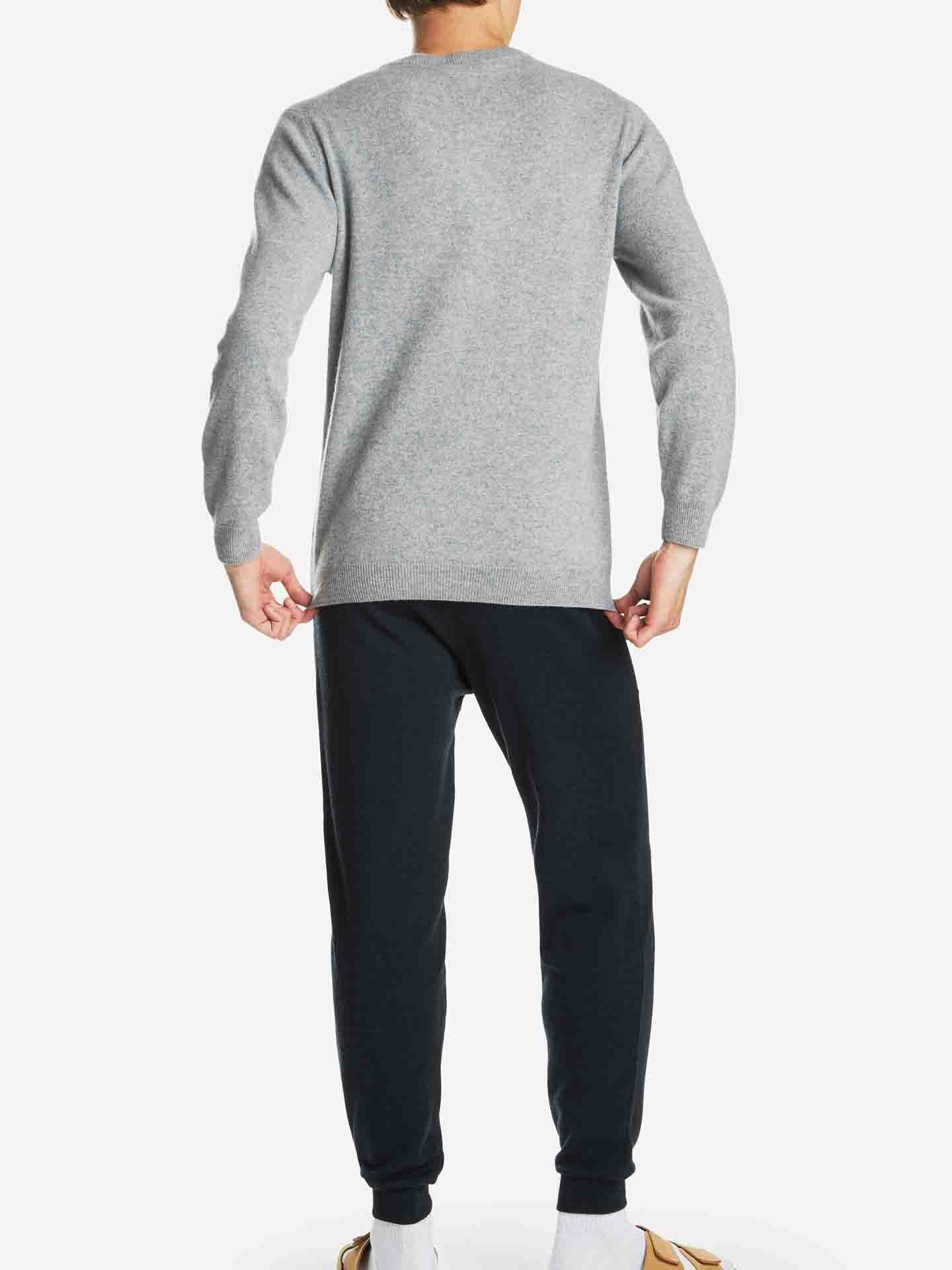 A person stands facing away, wearing a Blefjell Cashmere Crewneck Men by We Norwegians in light grey, dark trousers, and light-colored shoes with white socks. The plain white background emphasizes the luxury of the clothing.