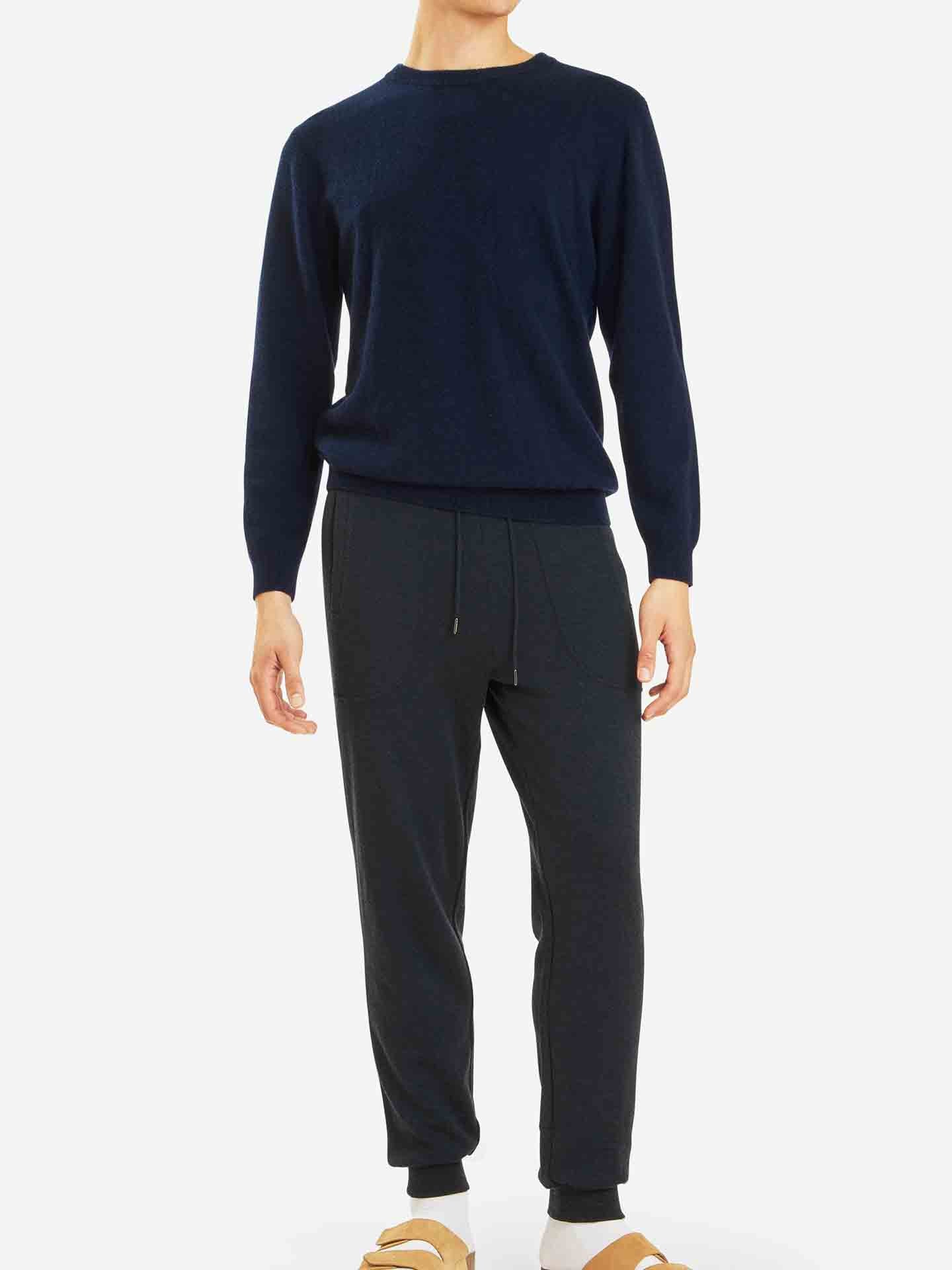 A person stands against a plain white background, donned in a dark blue Blefjell Cashmere Crewneck Men sweater by We Norwegians and black jogger pants. Beige slippers complete the outfit. The head is cropped out of the image.