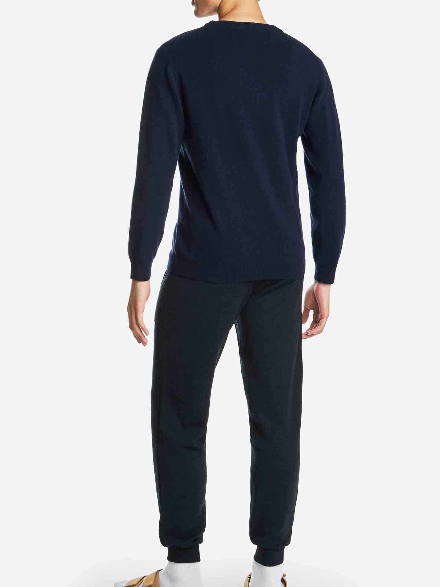 A person is seen from the back, wearing a luxurious Blefjell Cashmere Crewneck Men sweater by We Norwegians in a dark hue and matching dark pants. The outfit is complemented by light-colored socks and brown shoes. The background is plain white.