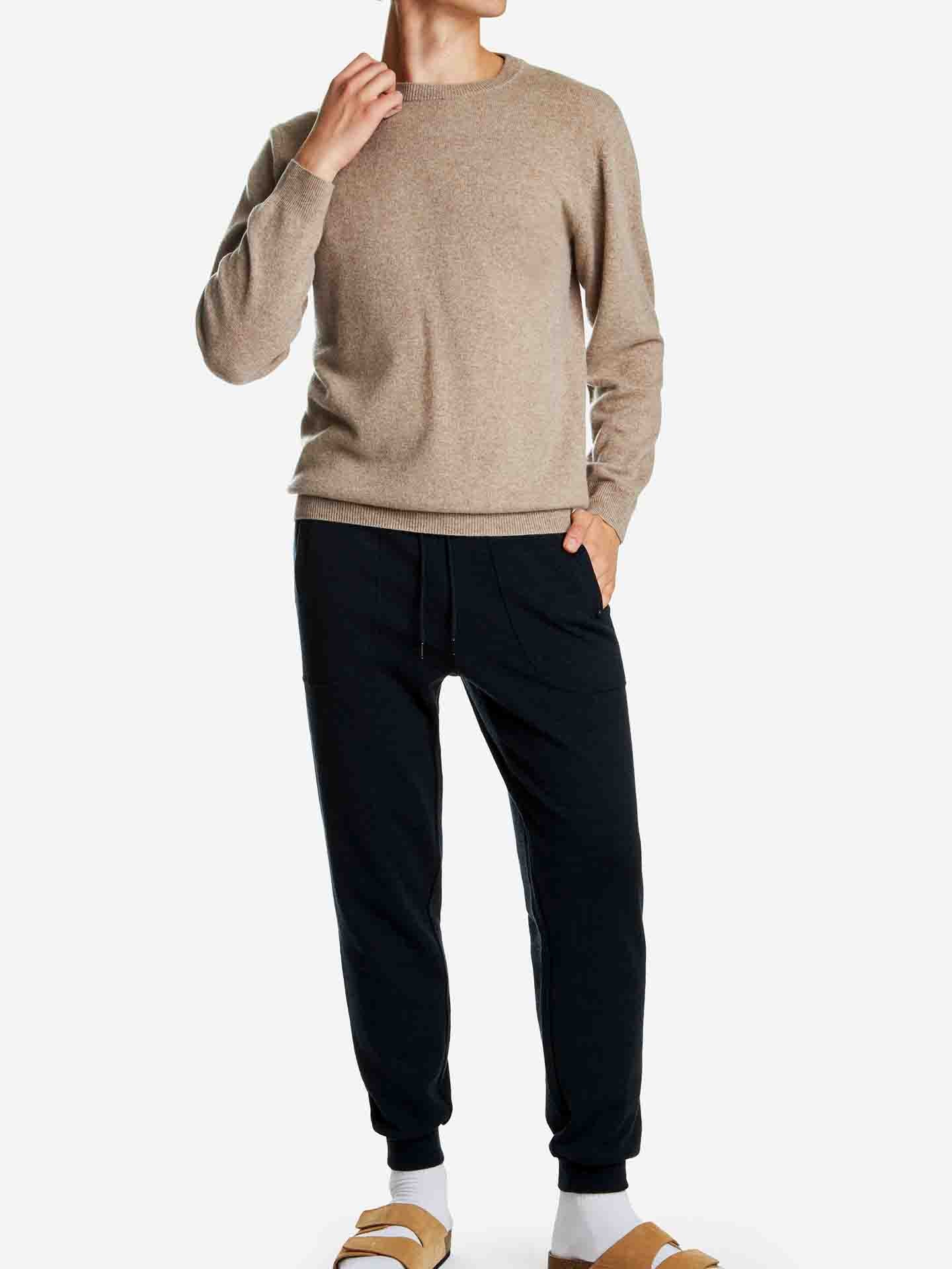 A person in a beige long-sleeve We Norwegians Blefjell Cashmere Crewneck Men, dark sweatpants, and beige sandals with white socks stands against a white background. One hand is tucked in a pocket, and the other touches the neck. The face is not visible.