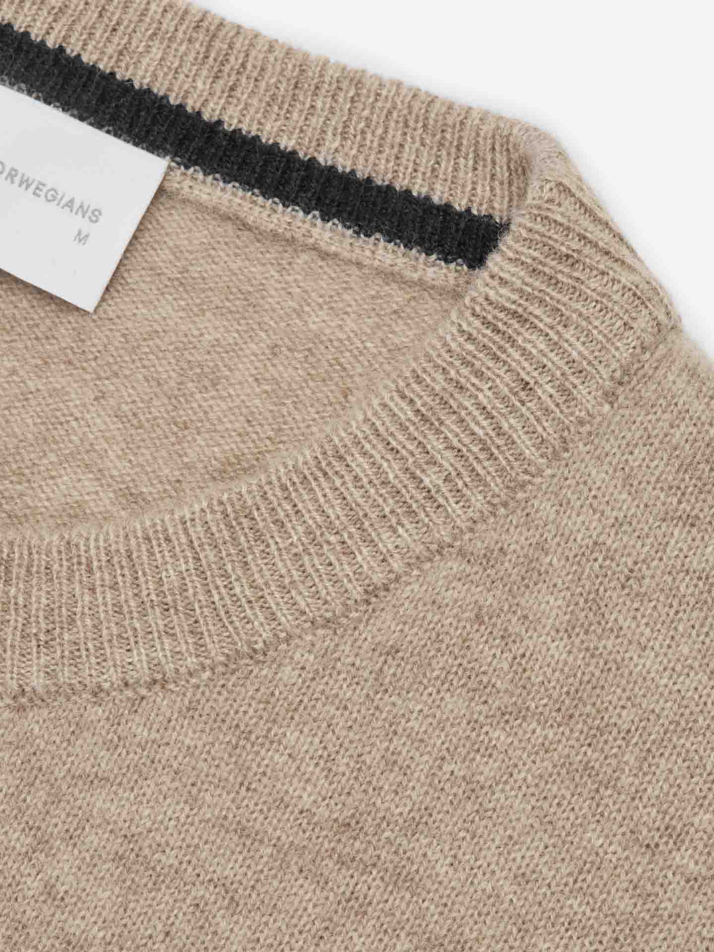 Close-up image of a luxury cashmere sweater's crew neck collar. The beige Blefjell Cashmere Crewneck Men features a ribbed neckline with a black stripe along the inside collar. A partially visible white label in the collar reads "We Norwegians" and "M.