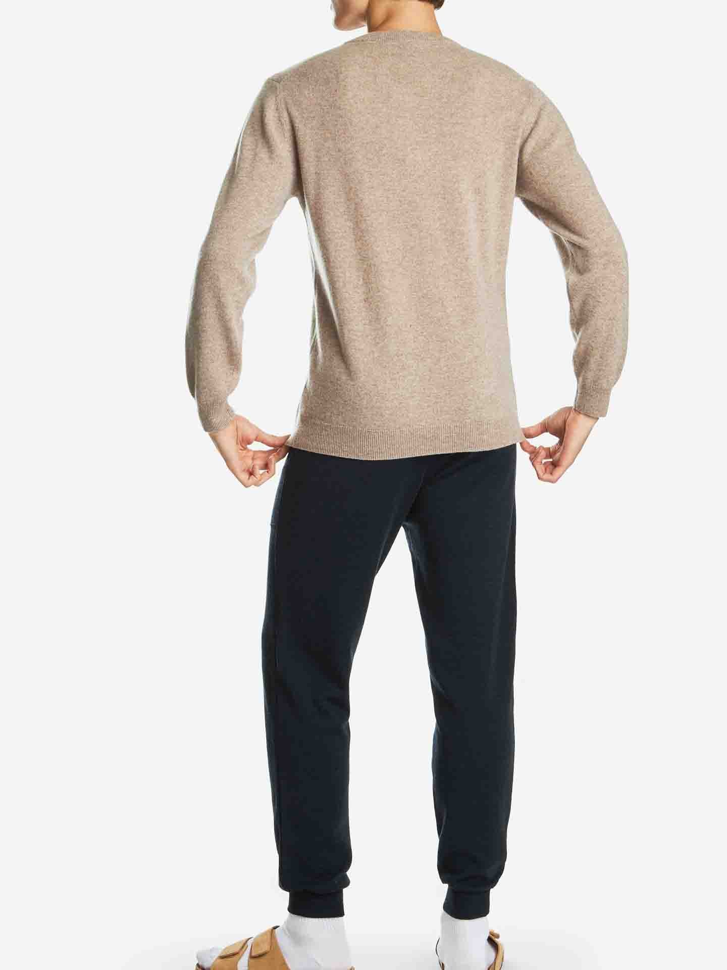 A person with short hair stands facing away from the camera, wearing a versatile luxury staple: the We Norwegians Blefjell Cashmere Crewneck Men in beige, paired with dark pants and light-colored sandals with white socks. The background is plain white.