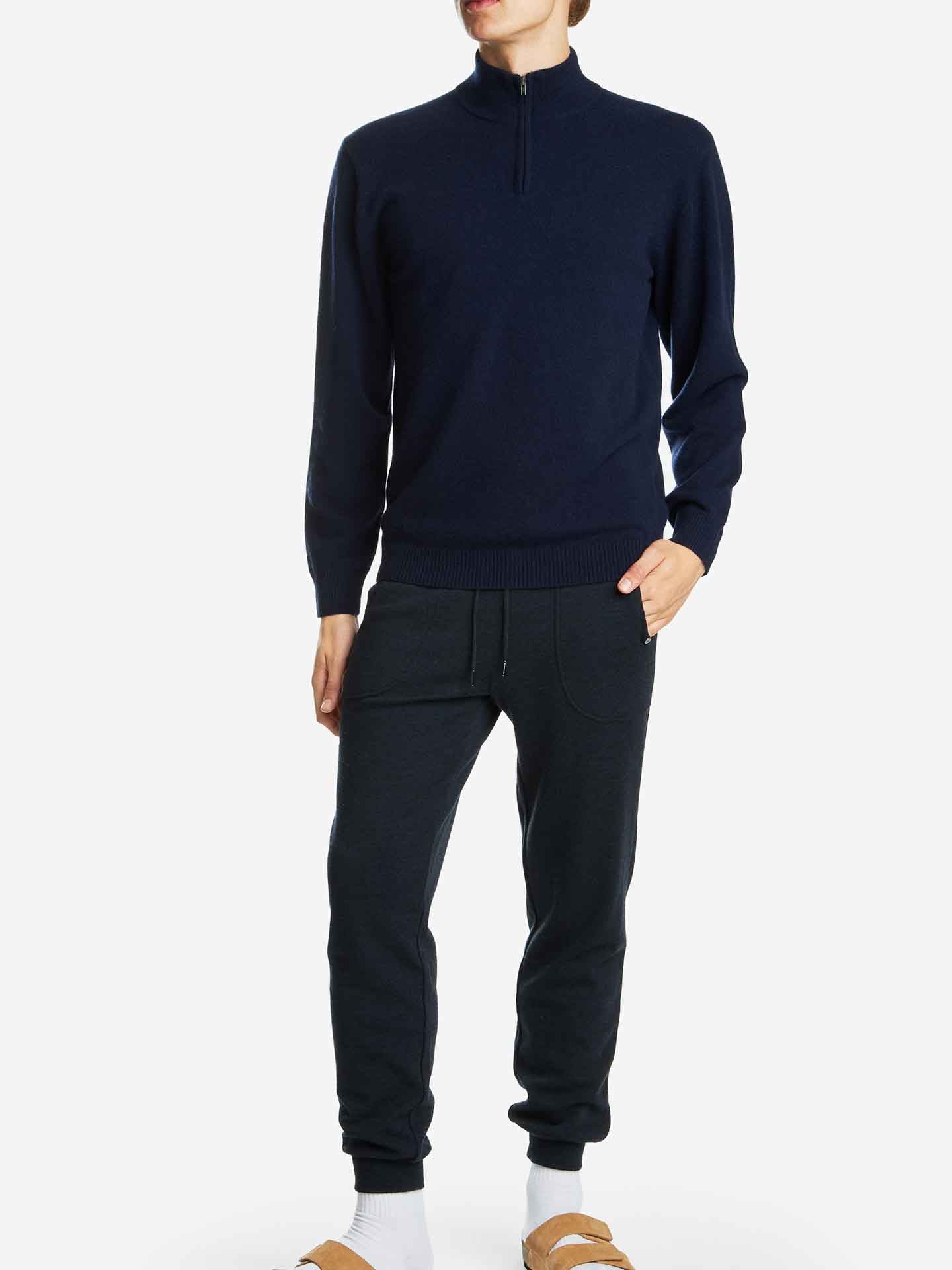 A person stands against a plain light background, wearing the Blefjell Zipup Men by We Norwegians, a dark navy cashmere sweater with a YKK zipper, and matching navy jogger pants. They also have on white socks and brown slip-on sandals. One hand is in their pocket.