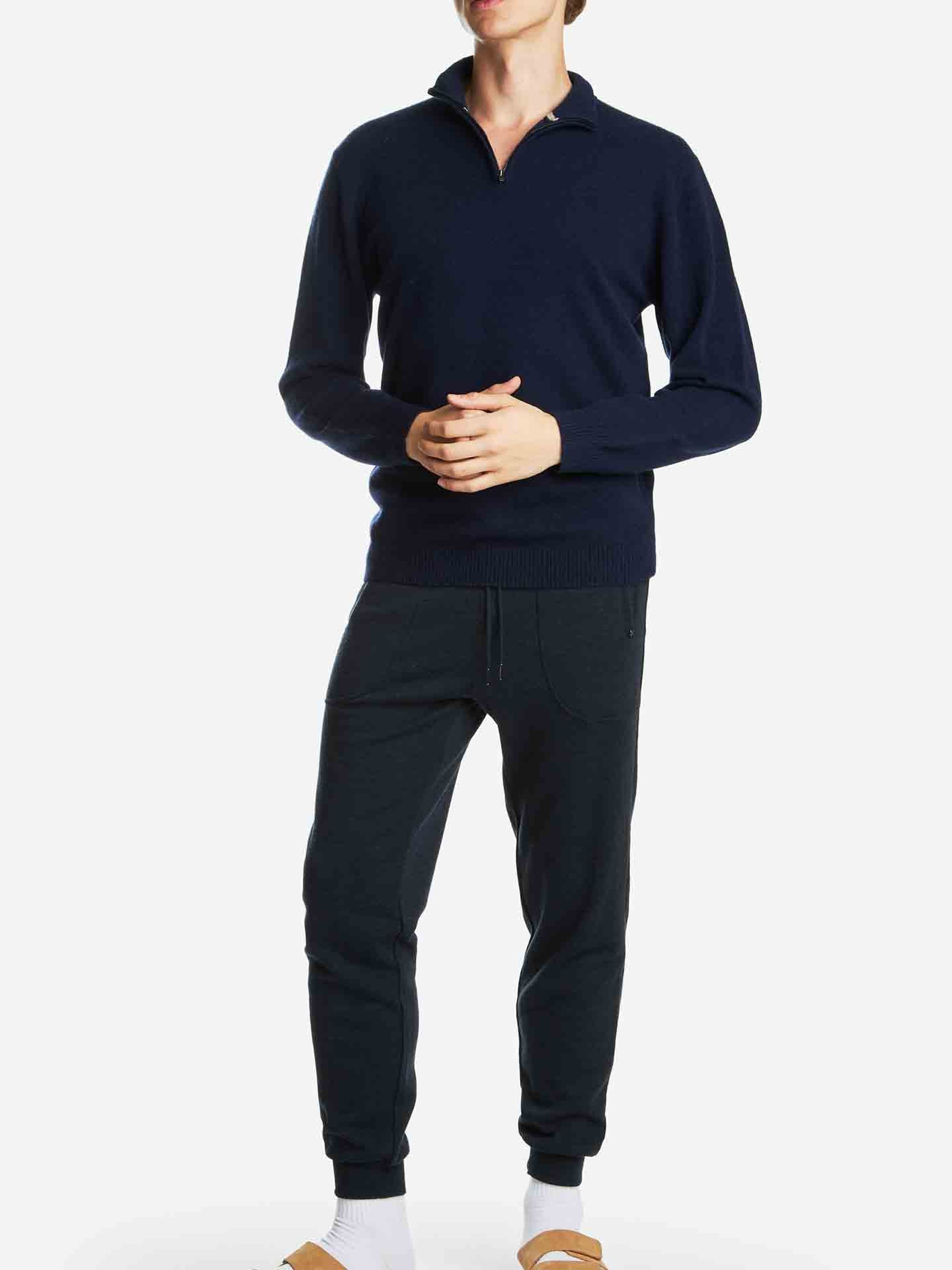 A person stands against a plain white background, wearing the luxurious Blefjell Zipup Men by We Norwegians, a navy blue cashmere quarter-zip sweater with a YKK zipper. They pair it with dark jogger pants, white socks, and tan slip-on shoes. Their hands are gently clasped in front of them, and their head is slightly tilted to the side.