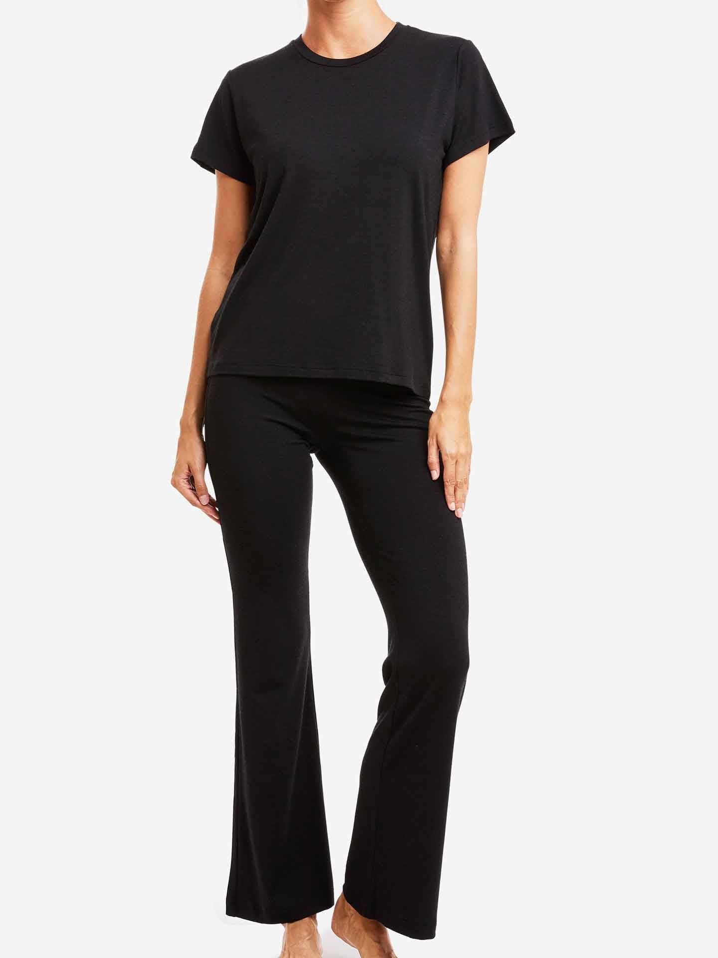 A person is dressed in a black Hygge Tee Women short-sleeved T-shirt, paired with flared pants from We Norwegians, crafted from soft Merino wool base layer material. They are standing against a white background, with their hands relaxed by their sides. The outfit appears comfortable and casual.