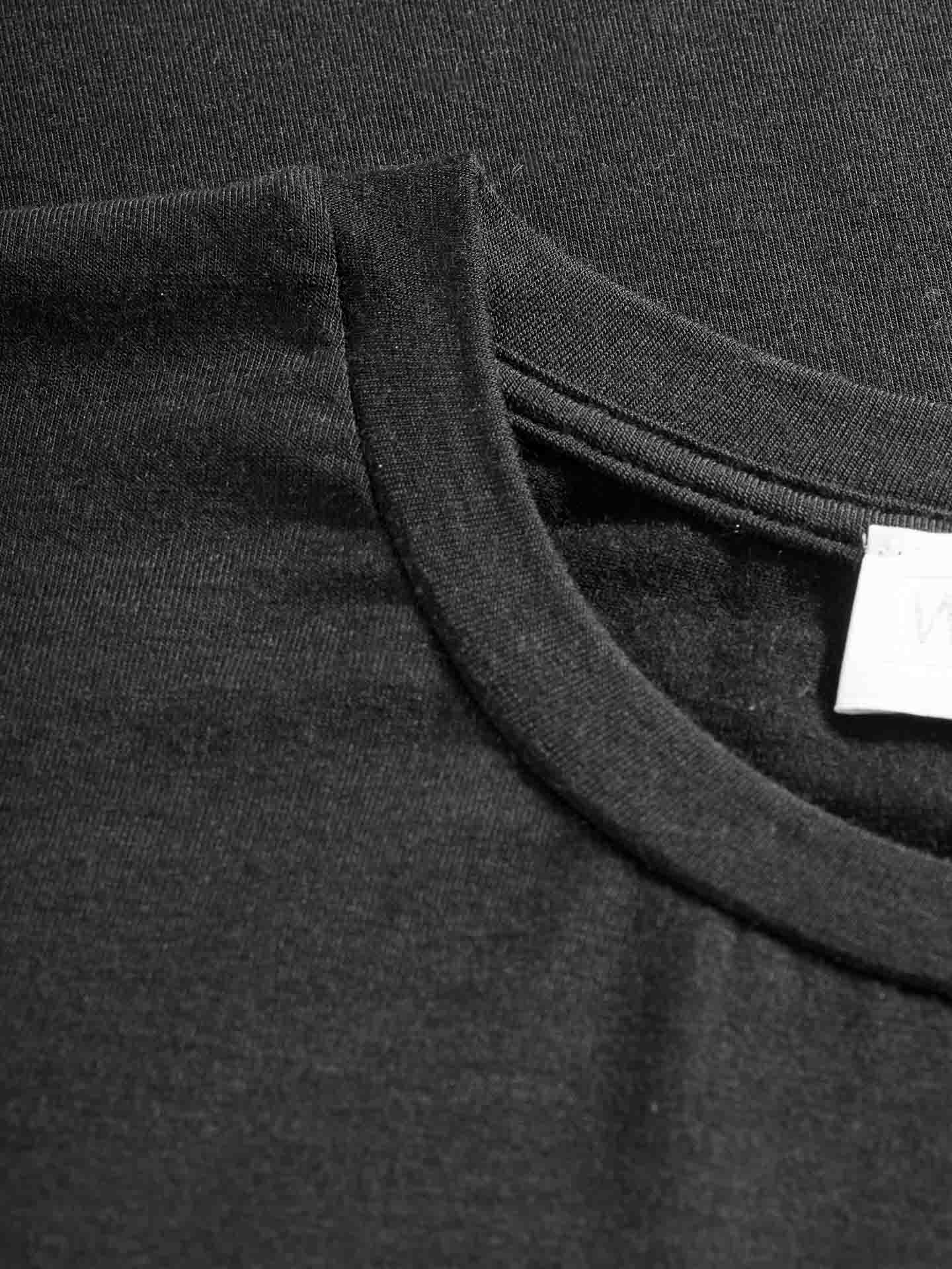 A close-up photograph of a black We Norwegians Hygge Tee Women base layer T-shirt, focusing on the collar and neckline. The fabric appears smooth, possibly merino wool, and the label can be partially seen, indicating the shirt's inside tag. The background is made from the same black material as the T-shirt, creating a seamless look.