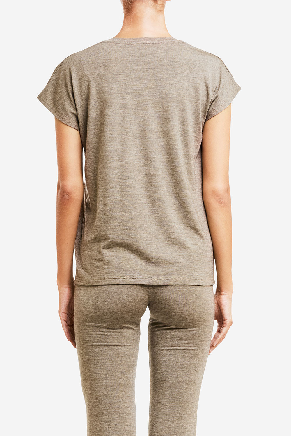 A person is standing with their back to the camera, wearing a light brown, short-sleeve "Hygge Tee Women" by We Norwegians and matching pants made from HYGGE base layer merino wool. The background is plain white. The person's hair is pulled back, and their right arm is slightly bent.