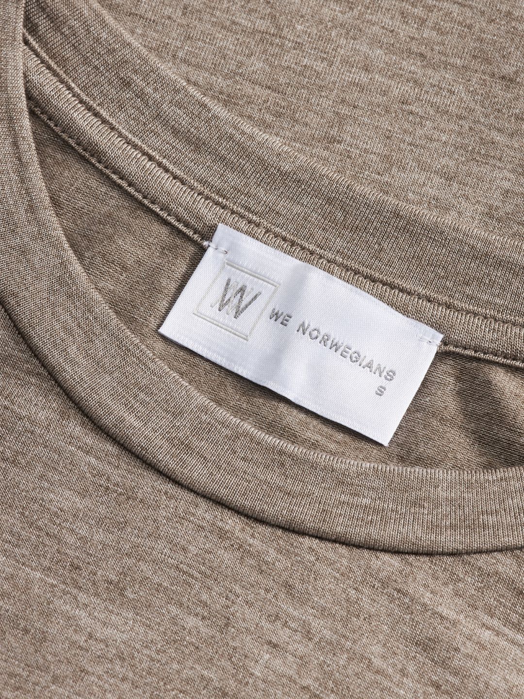 Close-up of a beige Hygge Tee Women luxury base layer with a white fabric label at the neck. The label shows a stylized "W" logo followed by the text "We Norwegians" and the size "S" printed below. The merino wool material has a smooth, fine-knit texture.