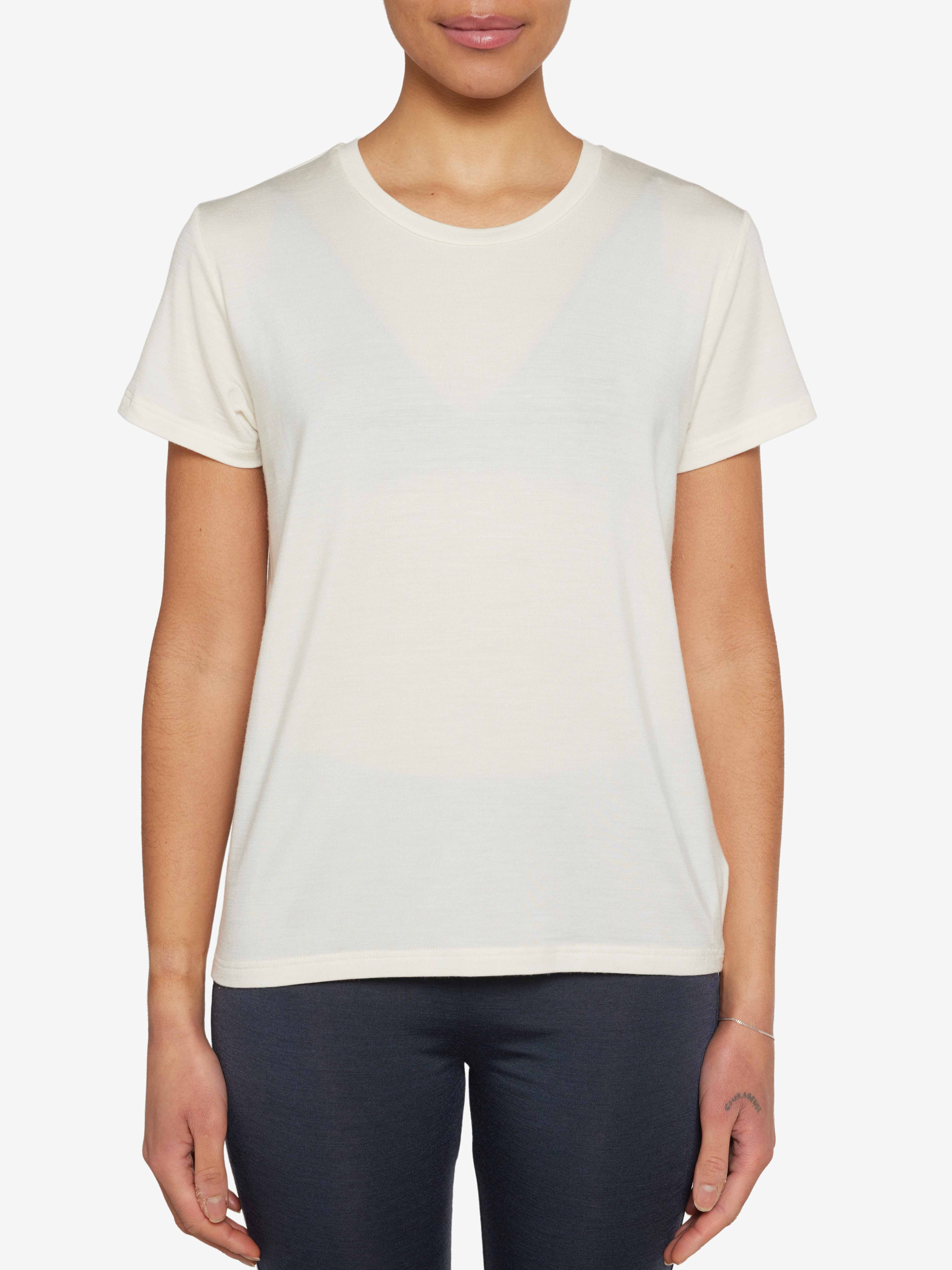 A person is standing against a white background, wearing a plain off-white Hygge Tee Women short-sleeve t-shirt from We Norwegians and dark pants. Their arms are relaxed at their sides, and only the upper torso and part of the legs are visible in the frame.