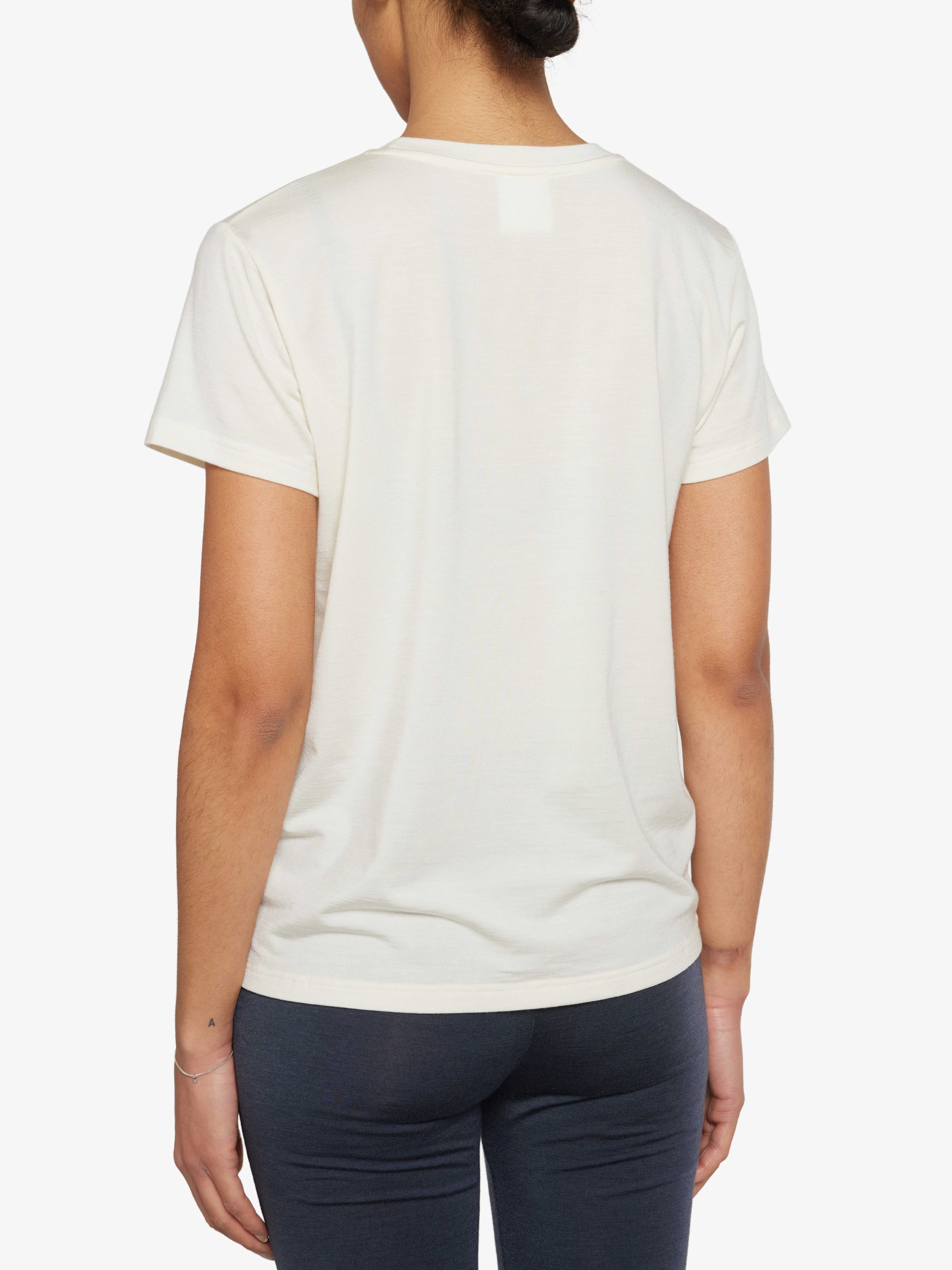 A person is standing with their back to the camera, wearing a plain light beige luxury base layer T-shirt called the Hygge Tee Women by We Norwegians and dark pants. Their arms are relaxed at their sides, and they have a bracelet on their right wrist. The background is white.