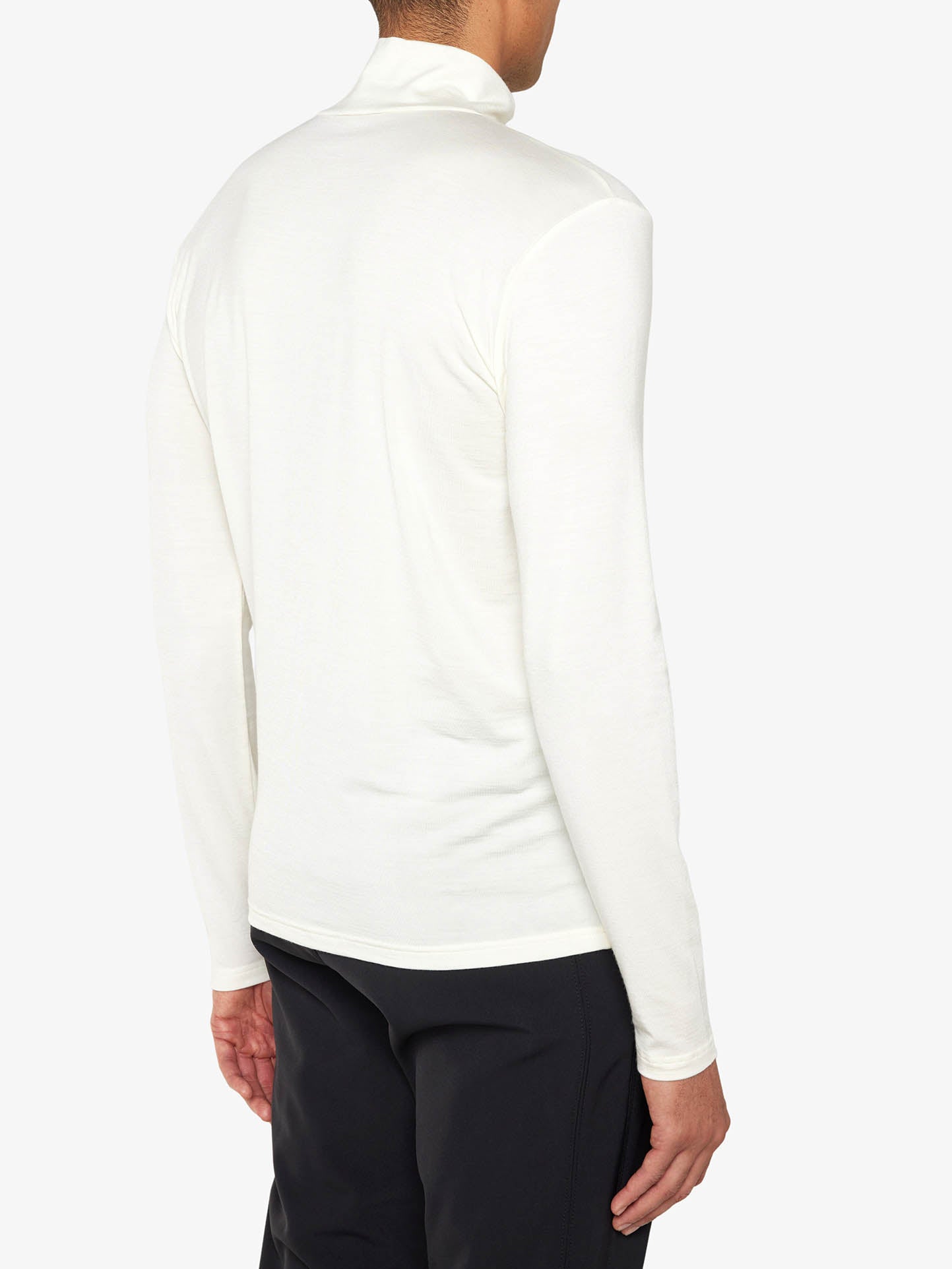 The image showcases an individual dressed in the "Hygge ZipUp Men" by We Norwegians, a white long-sleeve turtleneck shirt crafted from merino wool, paired with black pants. The photograph, taken from behind against a plain white backdrop, captures the slight turn of the person's head to reveal the high neckline of this versatile piece.