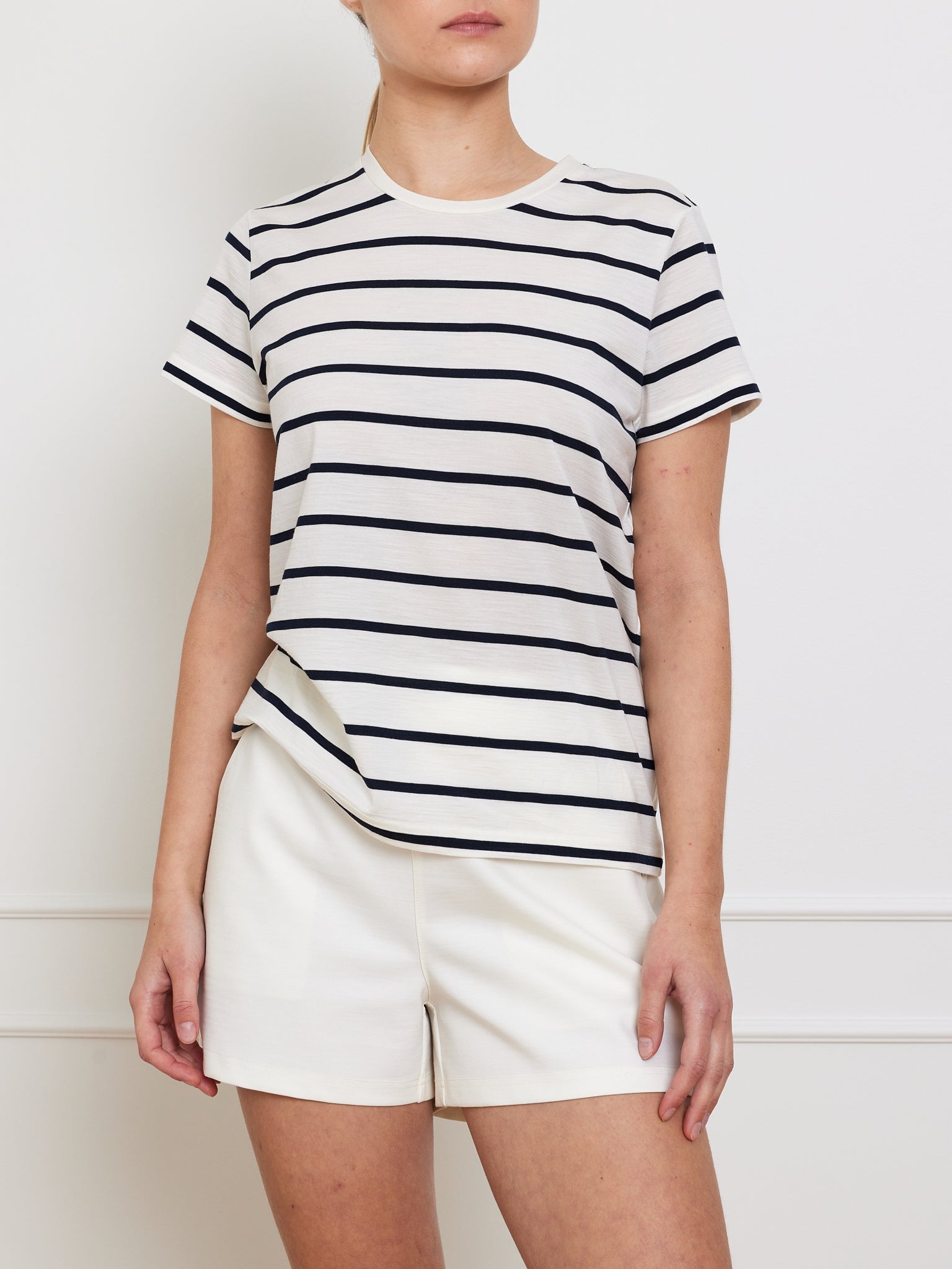 A person models the We Norwegians Kragero Tee Women White, a black and white striped short-sleeve T-shirt made of soft Merino wool, paired with white shorts against a plain backdrop, with their face not shown.