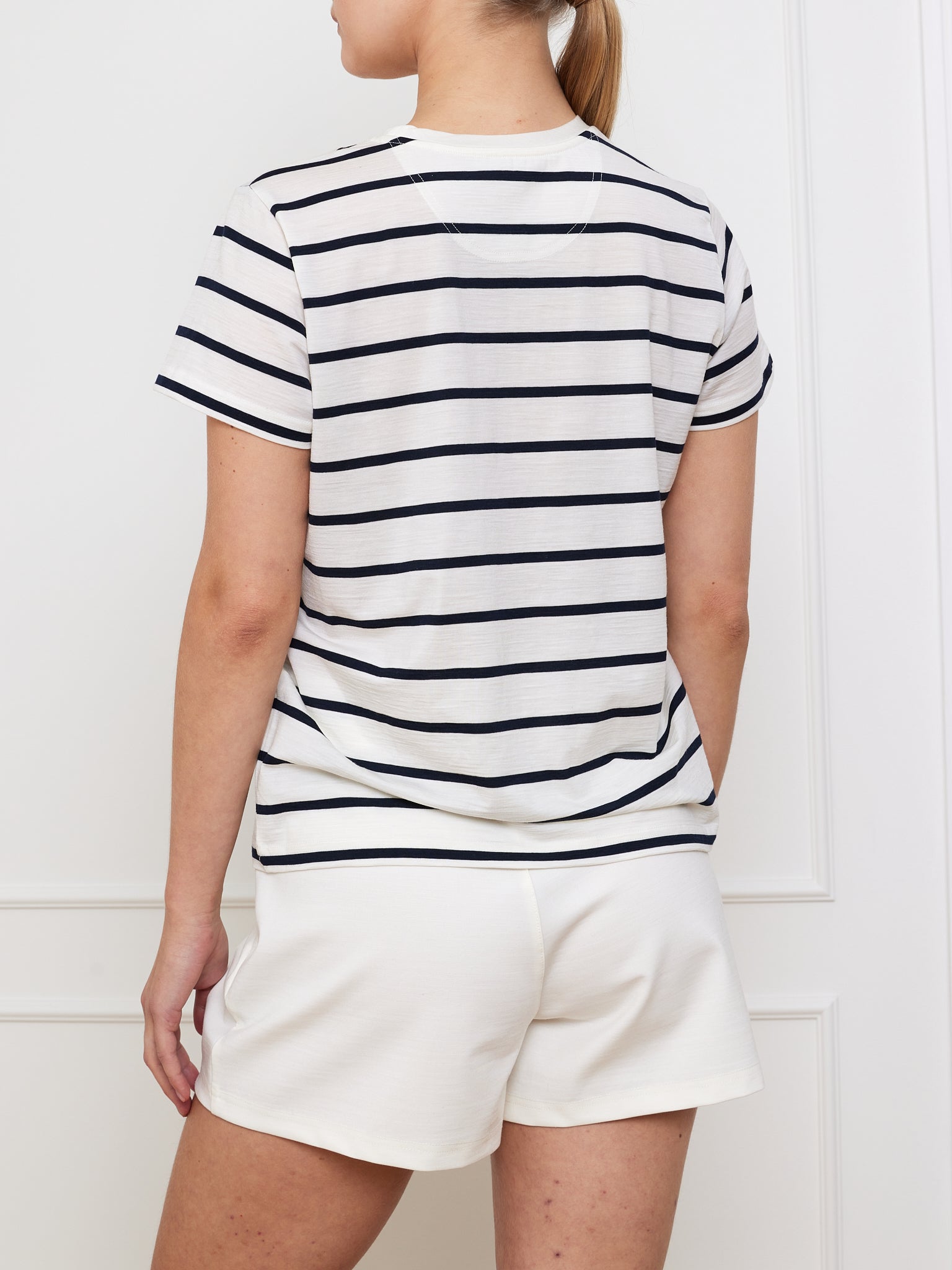 A person with a ponytail stands with their back to the camera, wearing the Kragero Tee Women White by We Norwegians, made from soft Tencel and breathable Merino Wool, paired with white shorts. The background is a white wall with paneling.