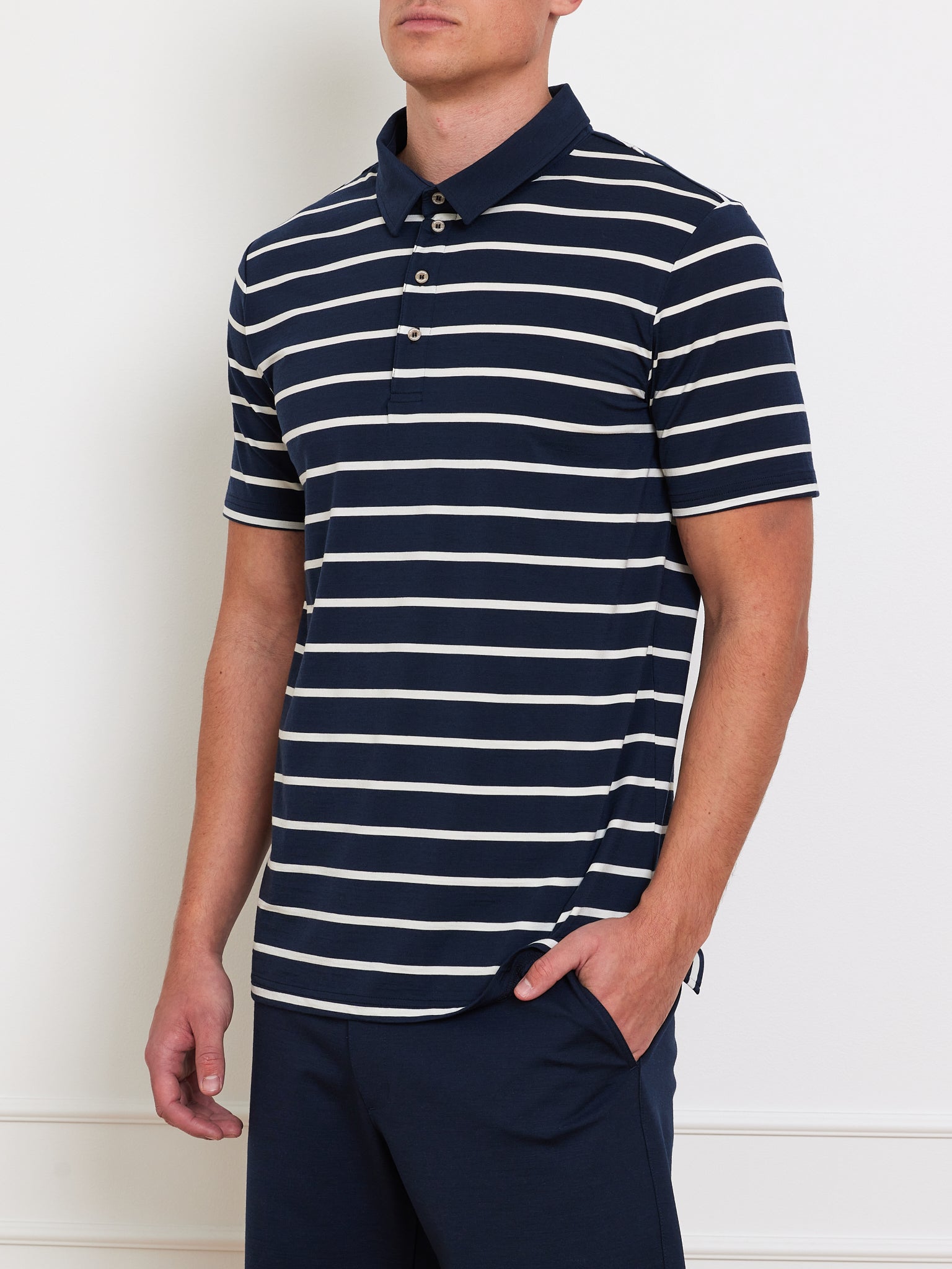 Sporting the Kragero Polo Men Navy by We Norwegians, a person creates a stylish spring/summer look with their left hand casually in their pocket against a plain, light-colored wall.