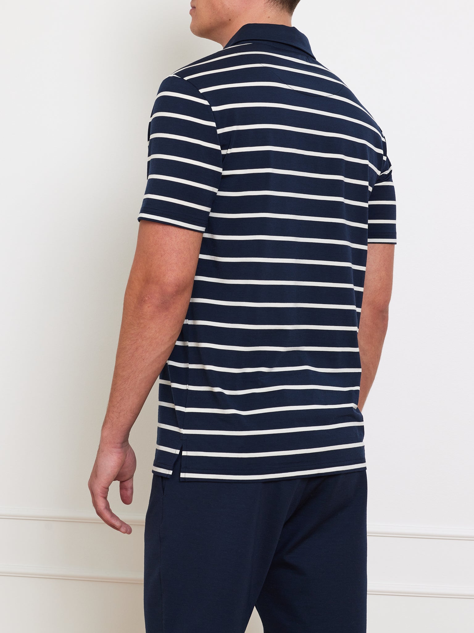 A person faces away wearing the Kragero Polo Men Navy from We Norwegians, a breathable navy blue and white striped Merino wool shirt, paired with navy pants. The short-sleeved, collared shirt suits spring/summer against a plain light wall backdrop.