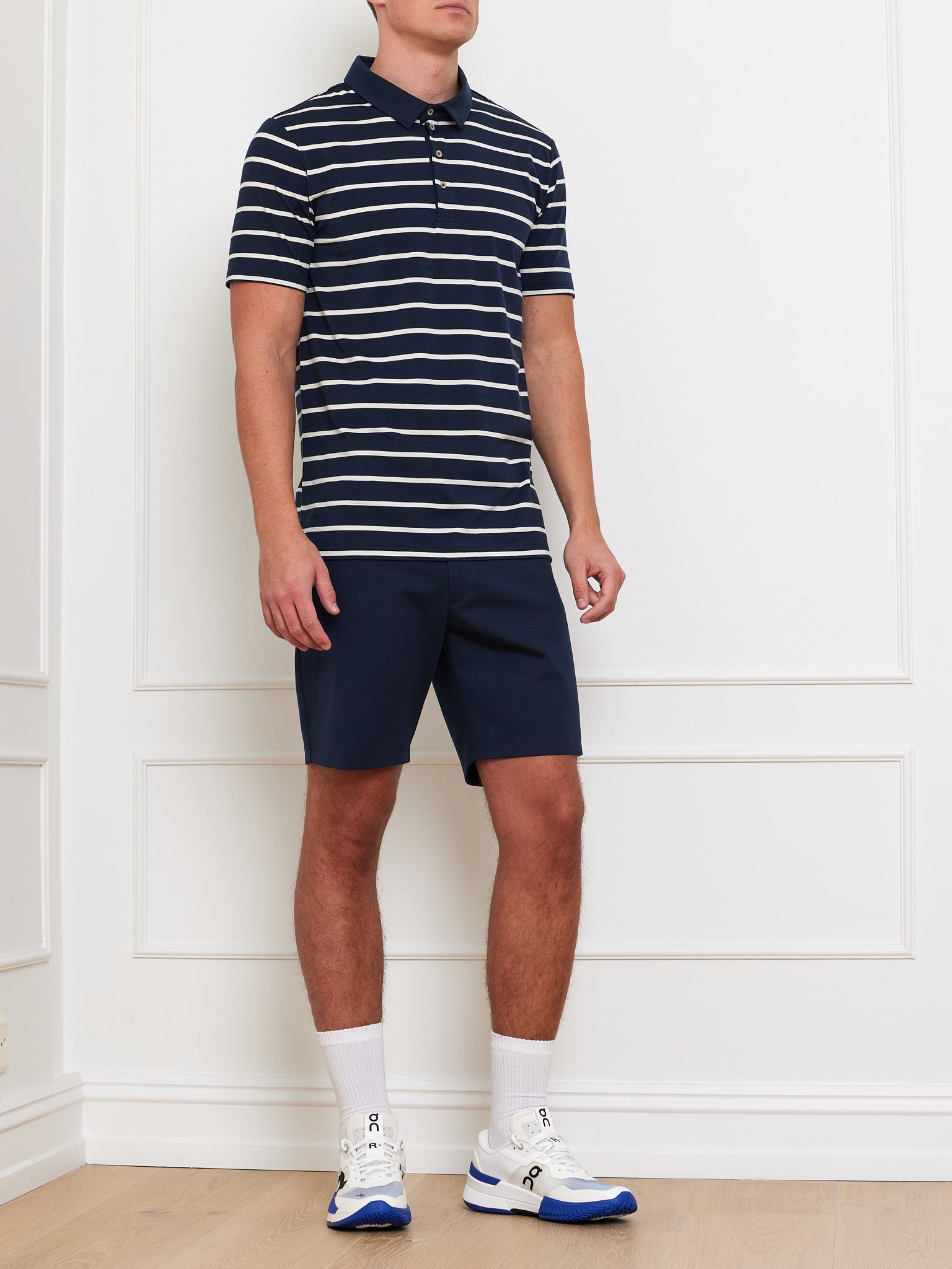 A person in a Kragero Polo Men Navy by We Norwegians, breathable navy shorts, white socks, and white sneakers with blue accents stands in a room with white paneled walls and light wood flooring. The head is out of view. Ideal for the Spring/Summer season.