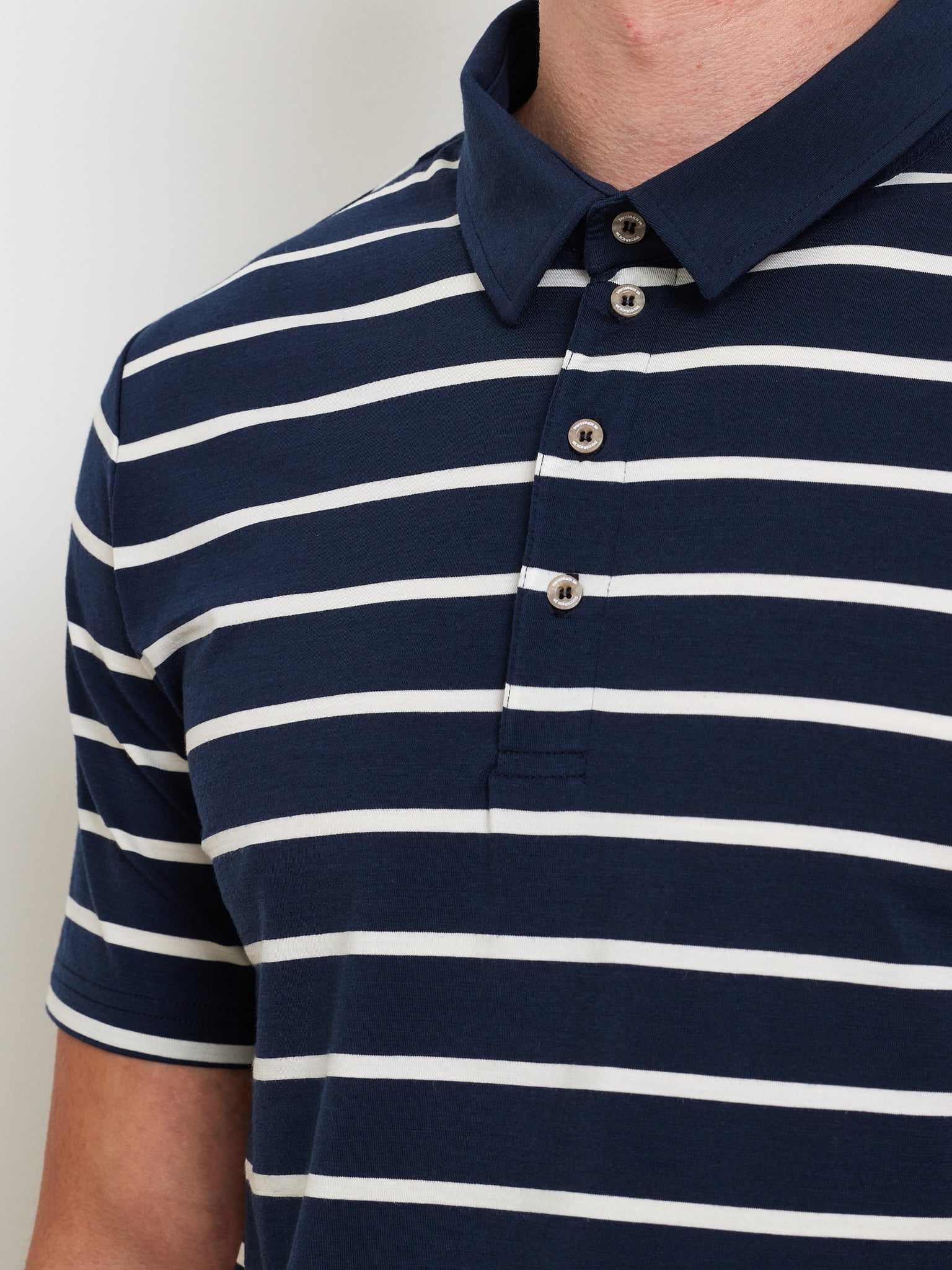 A close-up of someone wearing the Kragero Polo Men Navy by We Norwegians, showcasing its navy blue and white horizontal stripes with a collar and four-button front. Crafted from breathable Merino wool, the focus is on the shirt and upper torso against a neutral background.