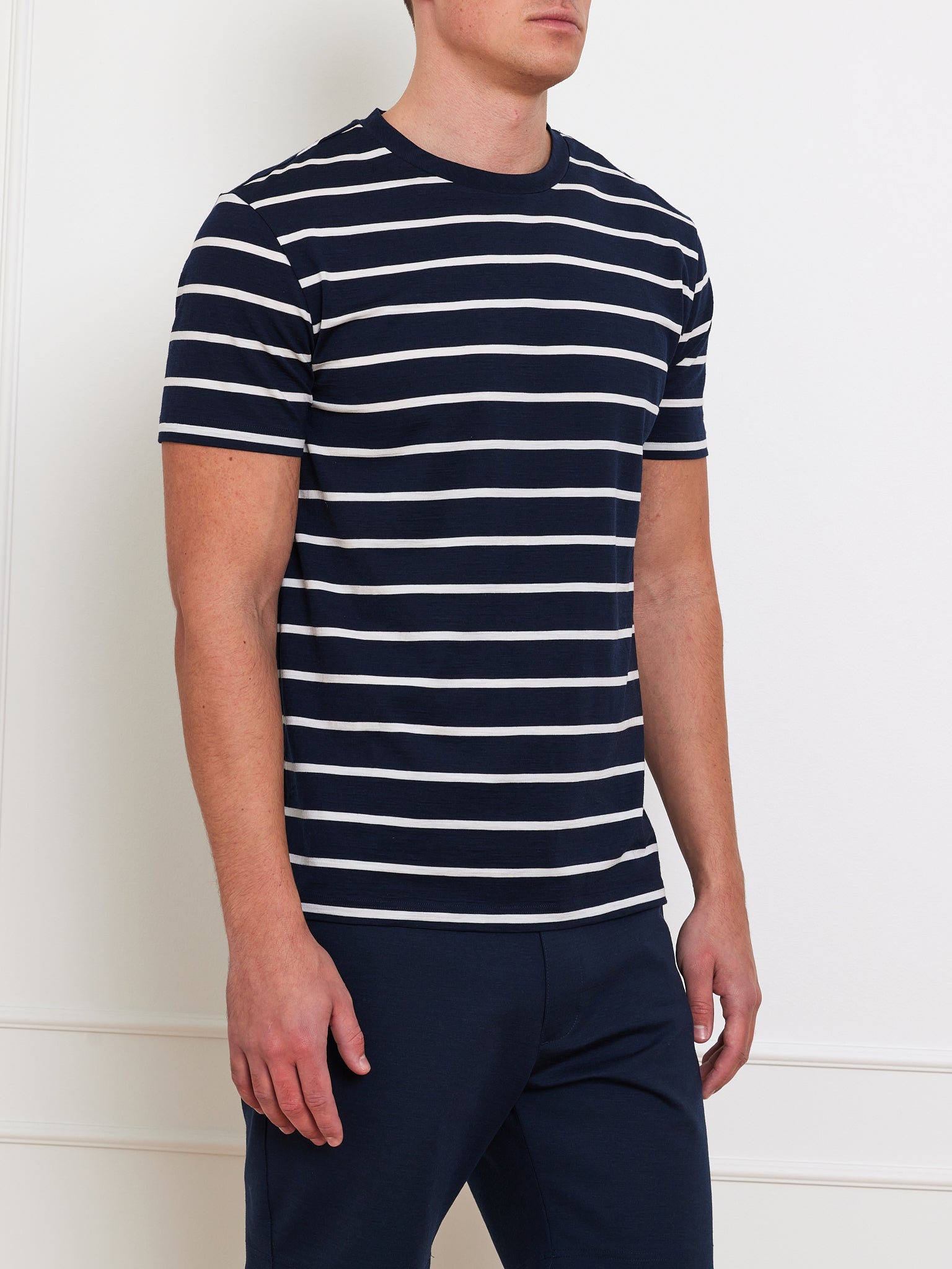 A person stands against a white wall wearing the We Norwegians Kragero Tee Men Navy, a breathable, navy blue and white striped crew neck t-shirt. The short-sleeved top pairs well with their dark pants. The person's face is not visible.