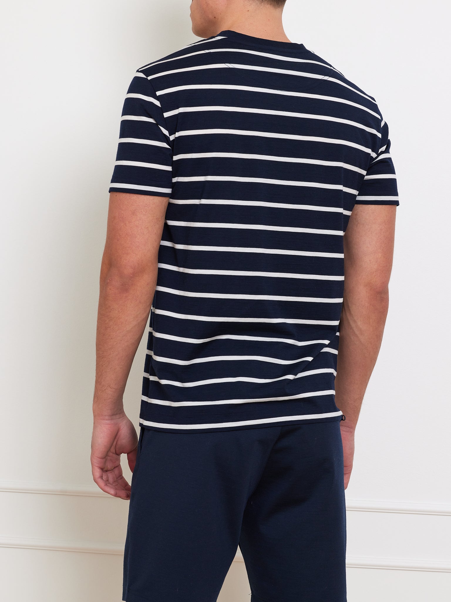 A person is wearing a We Norwegians Kragero Tee Men Navy, featuring white horizontal stripes, along with breathable navy blue pants. They are standing against a white background, facing away from the camera.