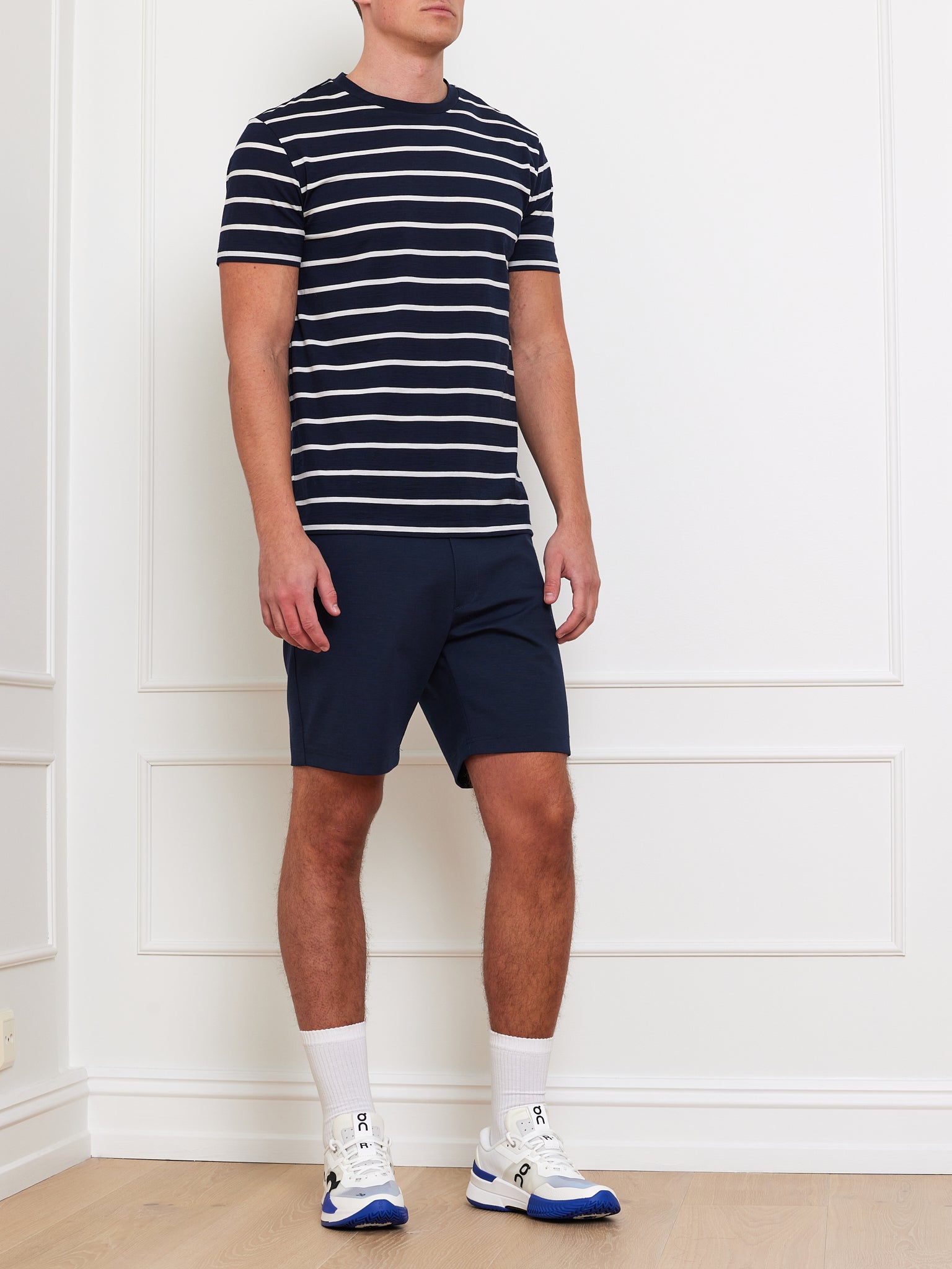 A man, with his face not visible, stands on a light wood floor against a white paneled wall, wearing the We Norwegians Kragero Tee Men Navy and navy shorts. White socks and sneakers with blue details complete his casual spring/summer look.