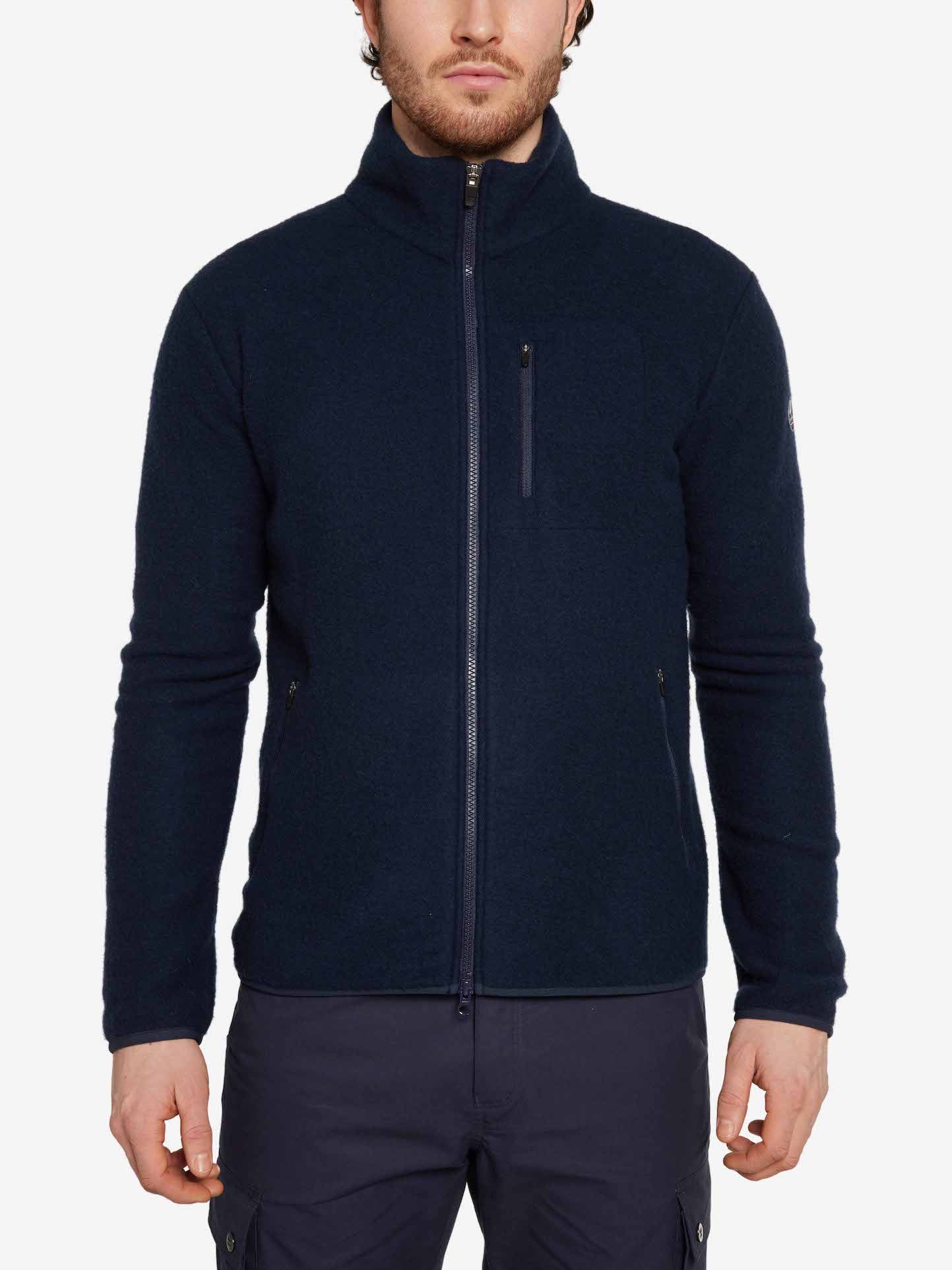 A person wearing the We Norwegians Svalbard Jacket Men in navy blue, a full-zip Merino wool fleece jacket featuring a high collar and zippered pockets, is shown from the waist up. The regular fit jacket is paired with dark blue pants. The background is plain white.