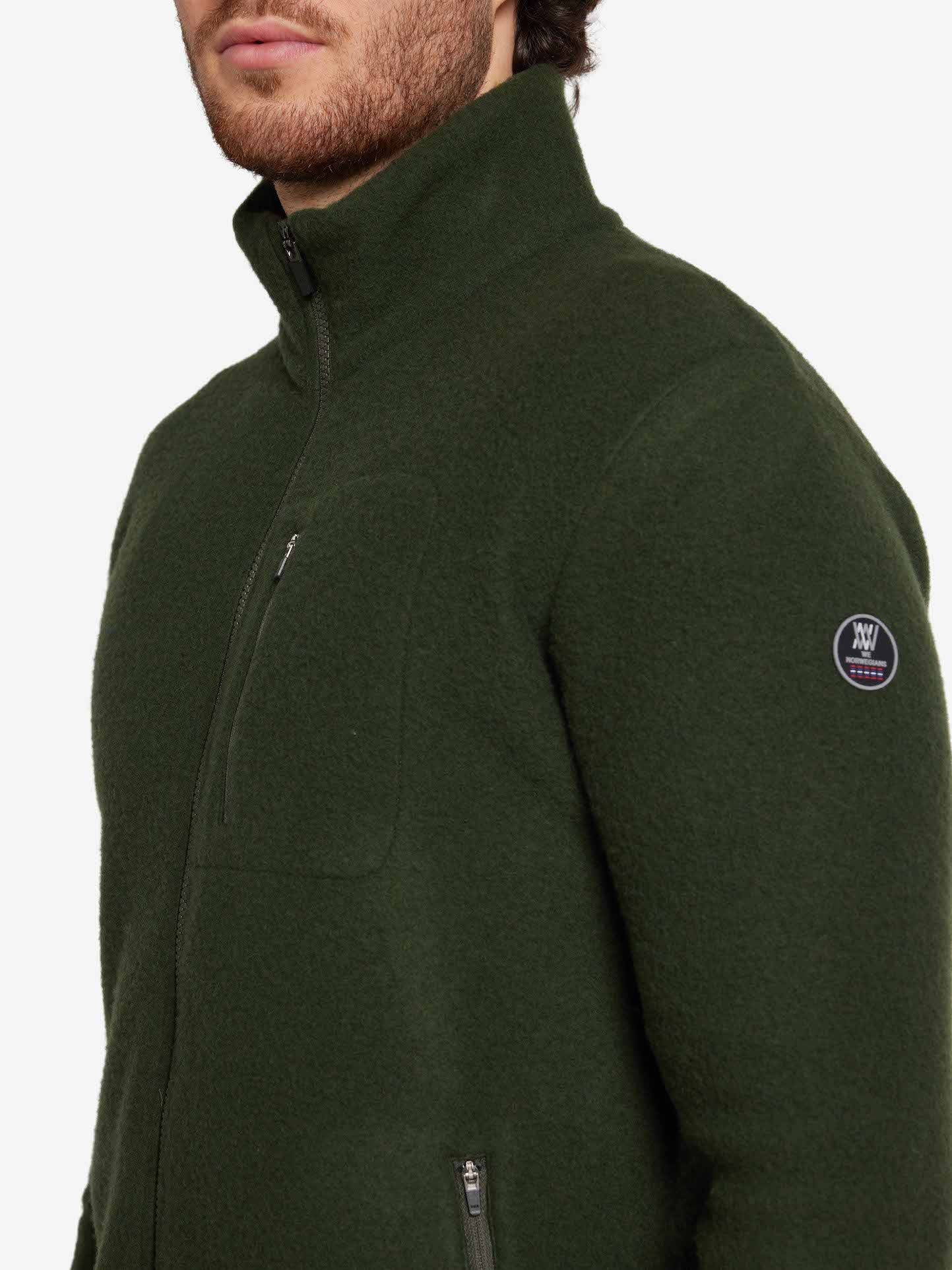 A person is wearing the We Norwegians Svalbard Jacket Men, a dark green, regular fit zip-up jacket with a high collar. The breathable jacket features a chest pocket and a round patch on the left sleeve. The person's face is partially visible against a plain white background.