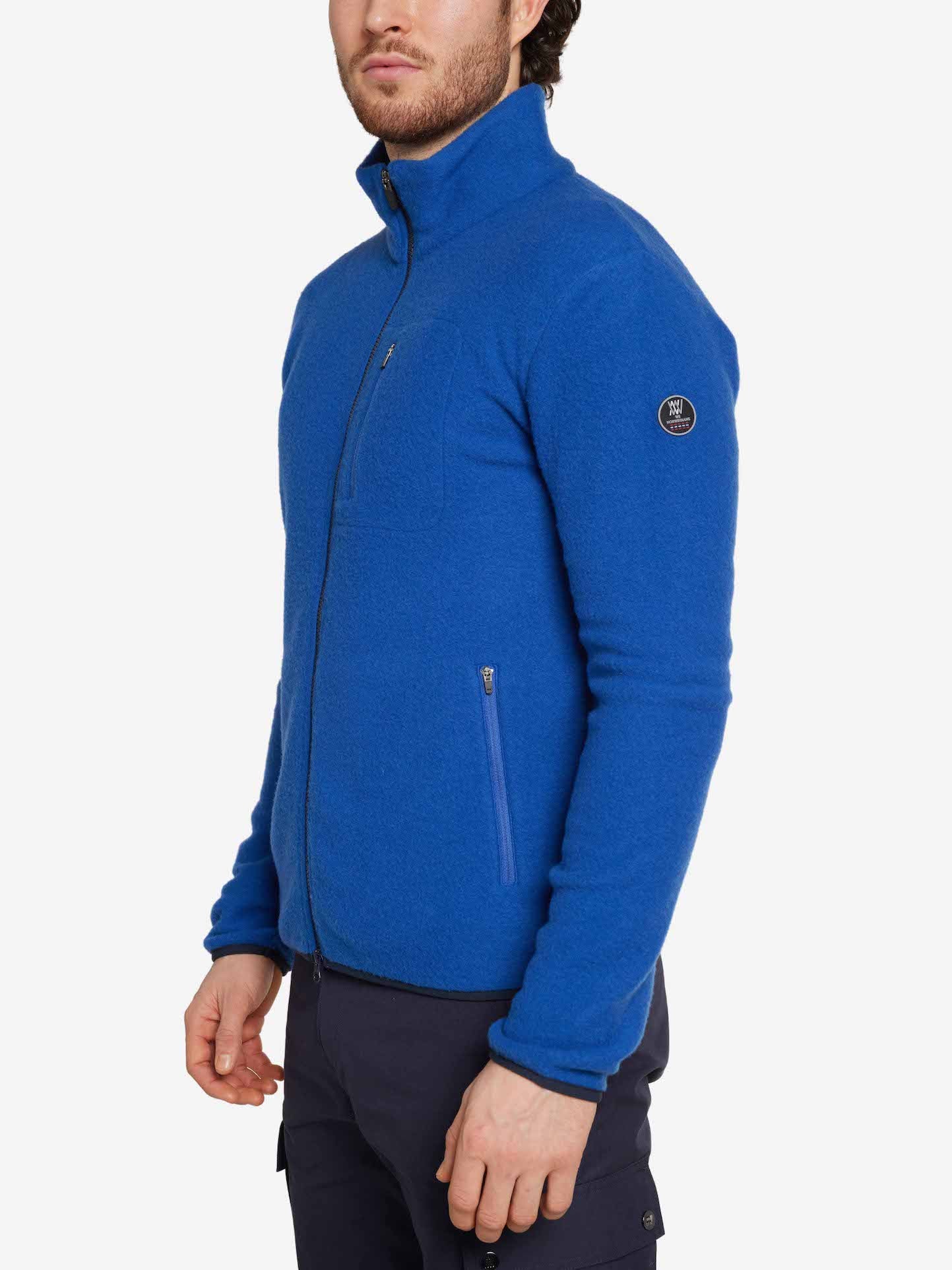 A person is standing and wearing the Svalbard Jacket Men by We Norwegians, a regular fit blue zip-up Merino wool fleece jacket with a small circular logo on the upper left sleeve. The jacket features a high collar and zippered pockets. They are also wearing dark blue pants. The background is plain white.