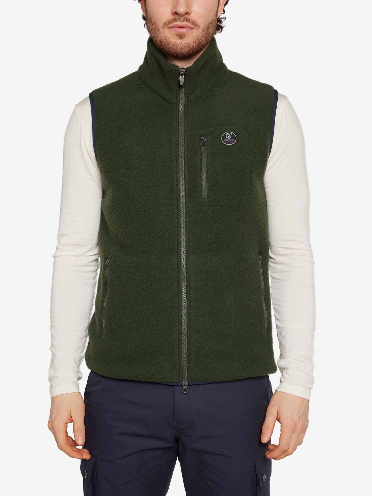 A person is wearing a Svalbard Vest Men by We Norwegians, featuring a dark green color and a zipper, over a white long-sleeve shirt and navy blue pants. The breathable vest, which has a small logo on the left side of the chest, offers a regular fit. The person's face is not visible.