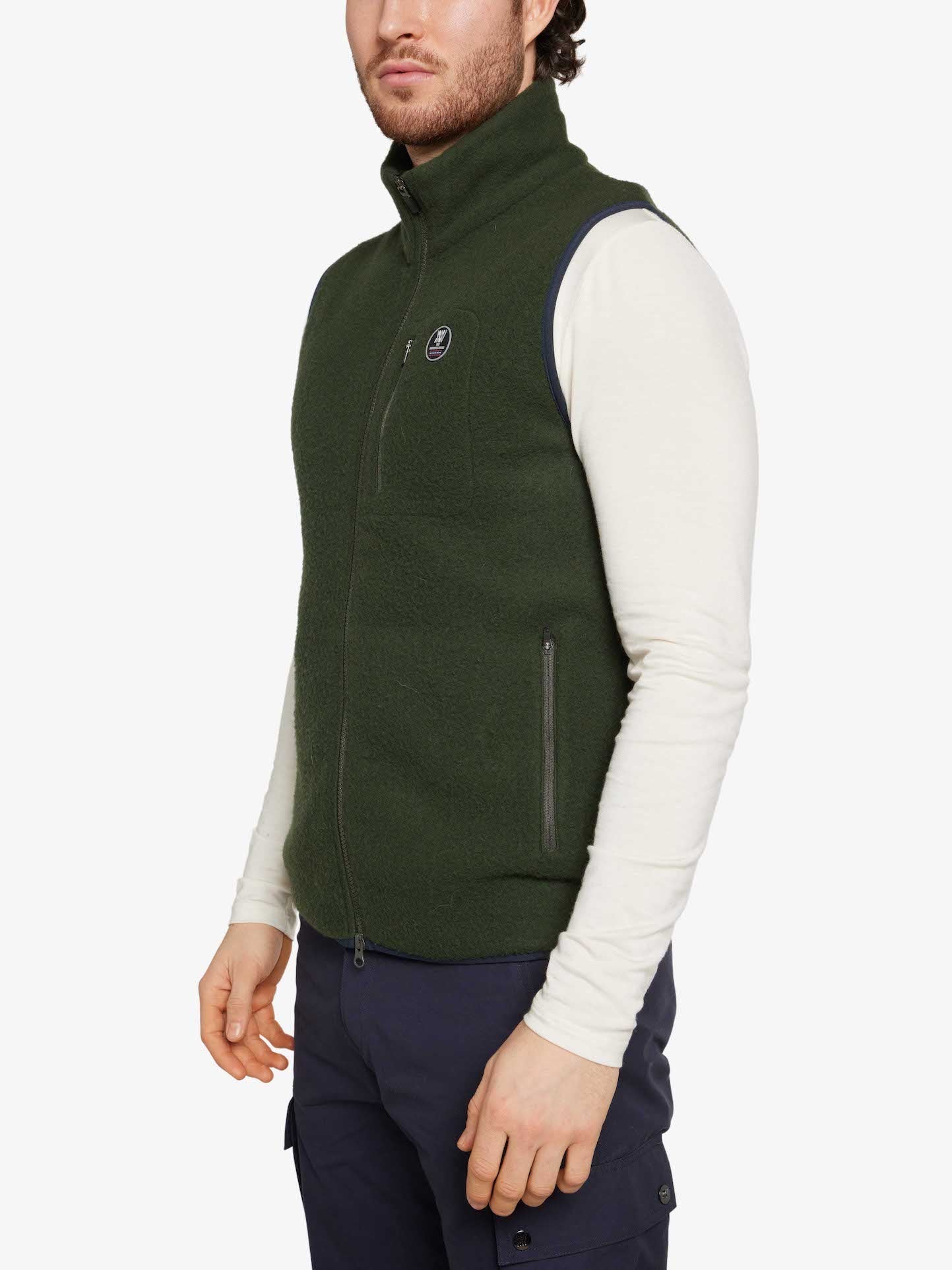 A person wearing a white long-sleeve shirt, the dark green We Norwegians Svalbard Vest Men, and navy cargo pants is standing against a plain white background. The regular fit vest features zippered pockets and a small circular logo on the left chest. Only the upper body and part of the legs are visible.