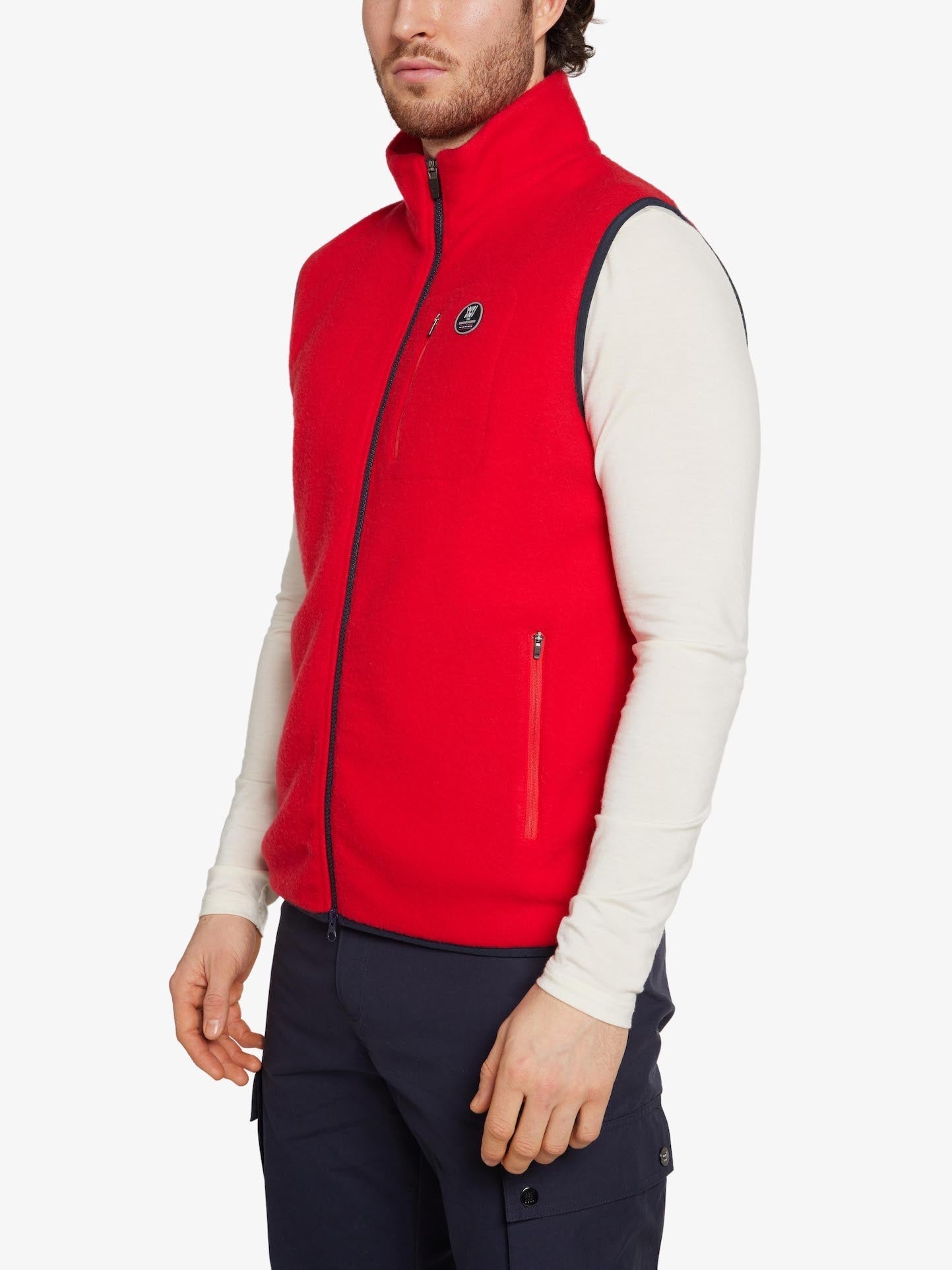 A person is wearing a red We Norwegians Svalbard Vest Men, made from breathable Merino fleece, over a long-sleeved white shirt and dark blue cargo pants. The regular fit vest features zippered pockets and a small emblem on the chest. The background is plain white.