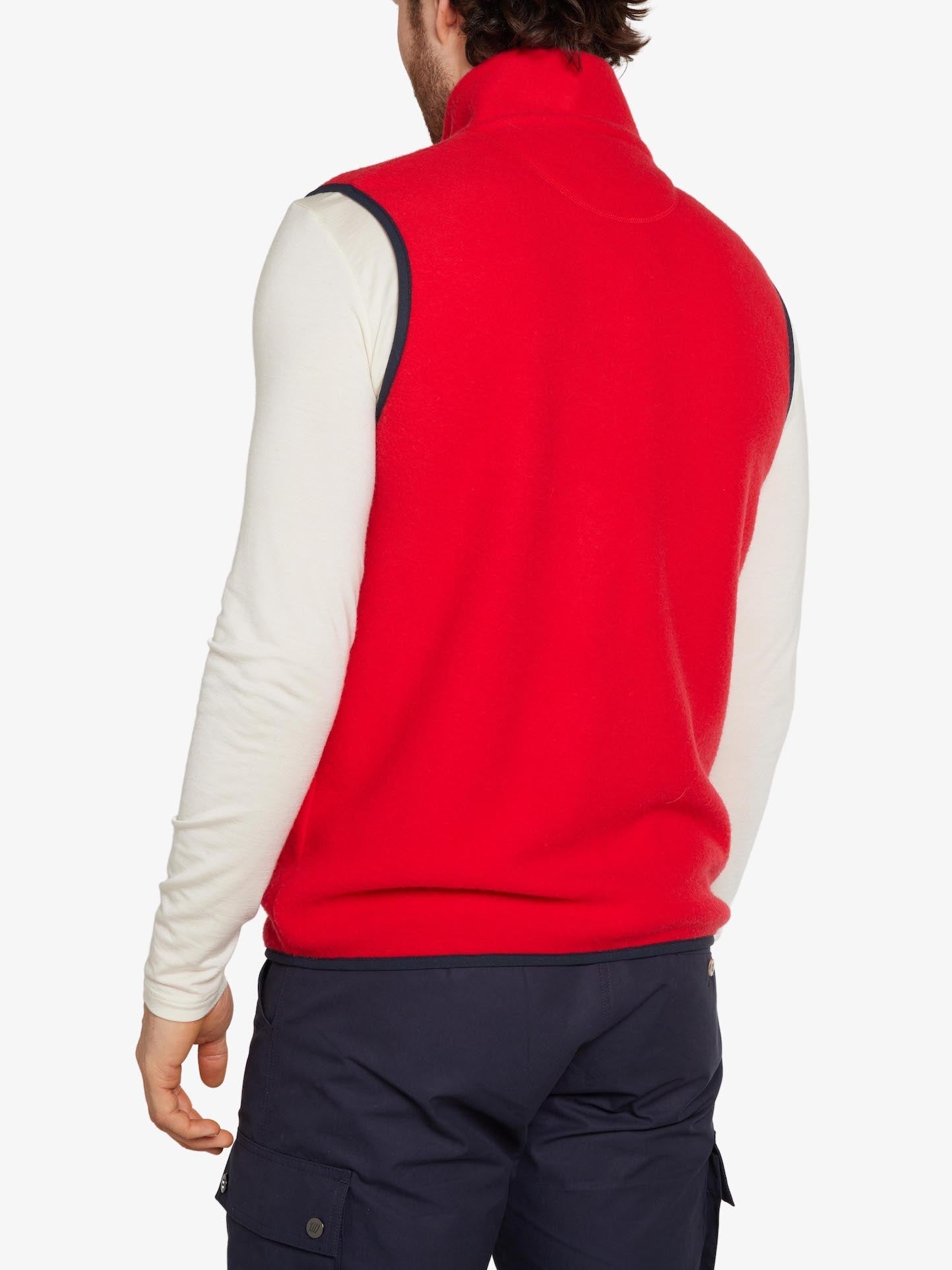 A man is seen from behind wearing a breathable Svalbard Vest Men by We Norwegians in red, layered over a long-sleeve white shirt and dark pants with multiple pockets. His head is turned slightly to the left against a plain white background.