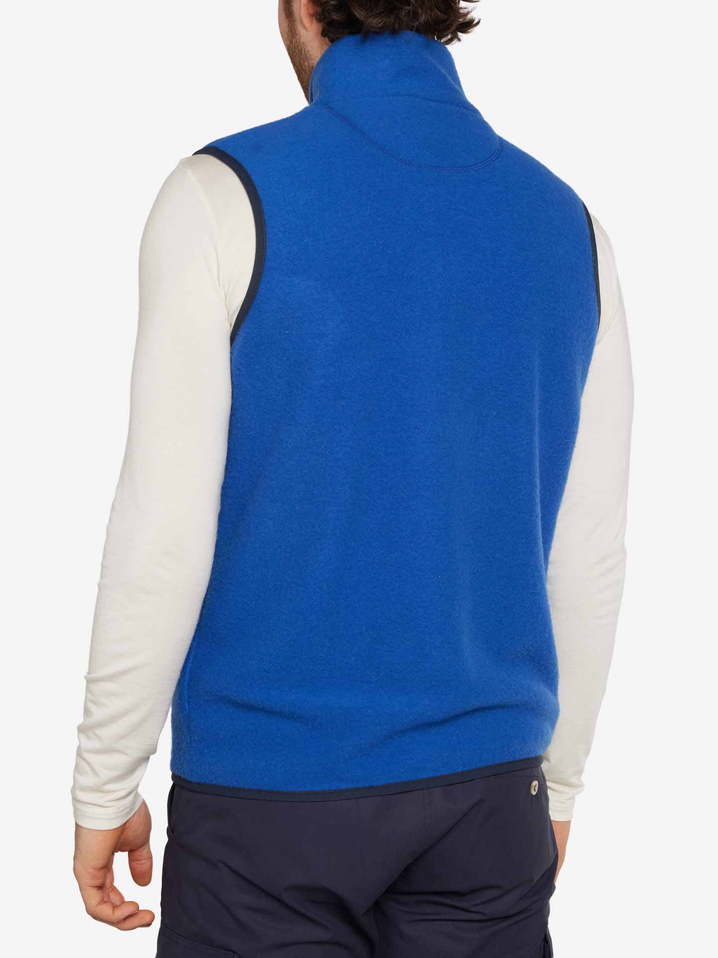 A person wearing the We Norwegians Svalbard Vest Men in bright blue, regular-fit Merino fleece over a white long-sleeve shirt and dark pants. Viewed from behind, the sleeveless design and high collar of the breathable vest are showcased.