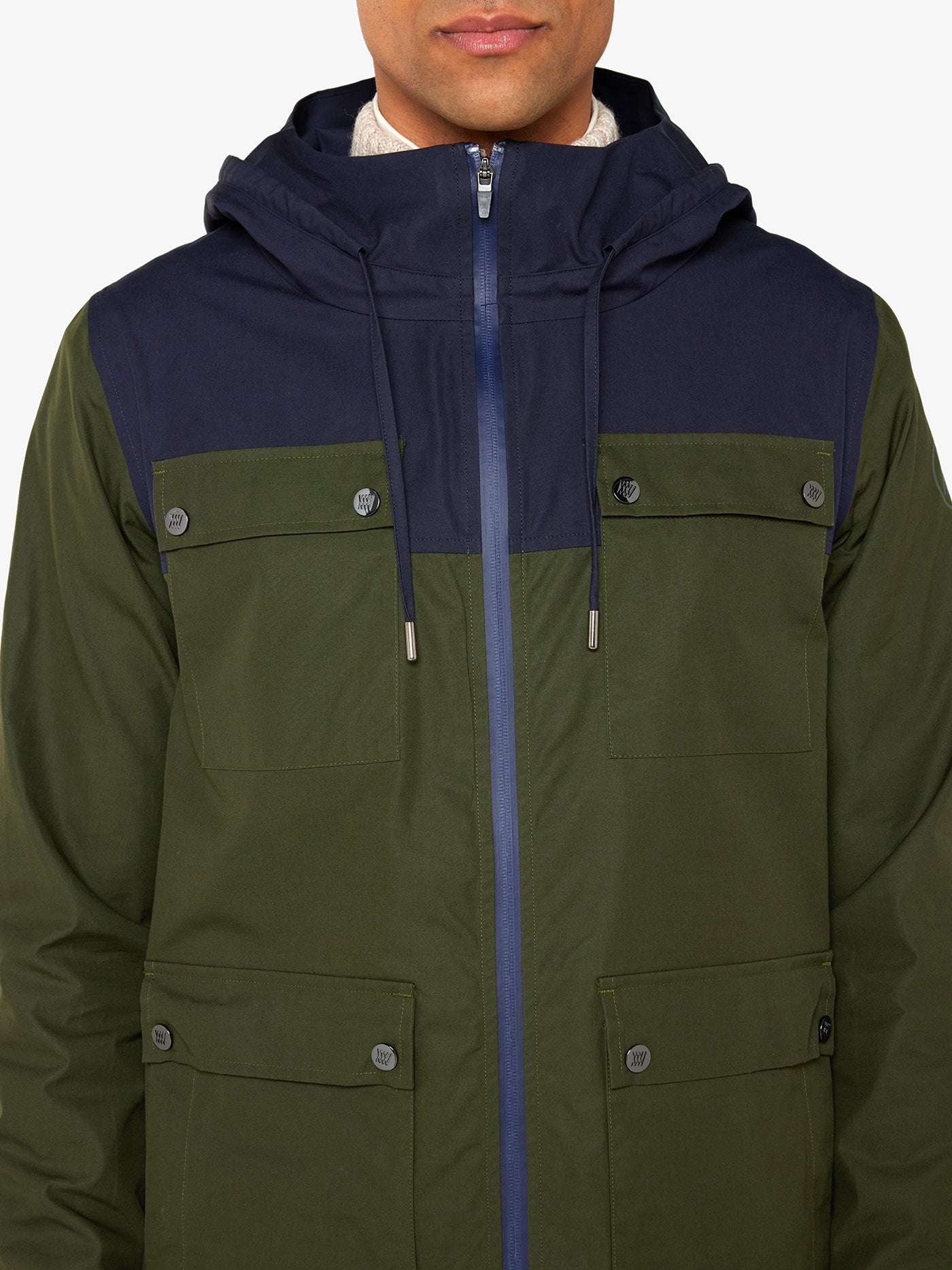 A person is wearing the We Norwegians Lyngen Jacket Men in olive green, crafted from premium Ventile® fabric. The jacket showcases a stylish two-toned design with a dark blue hood and chest area while the lower sections are in olive green. Ideal for outdoor enthusiasts, it features a front zipper, buttoned pockets on the chest, and flap pockets on the sides. The background is plain white.