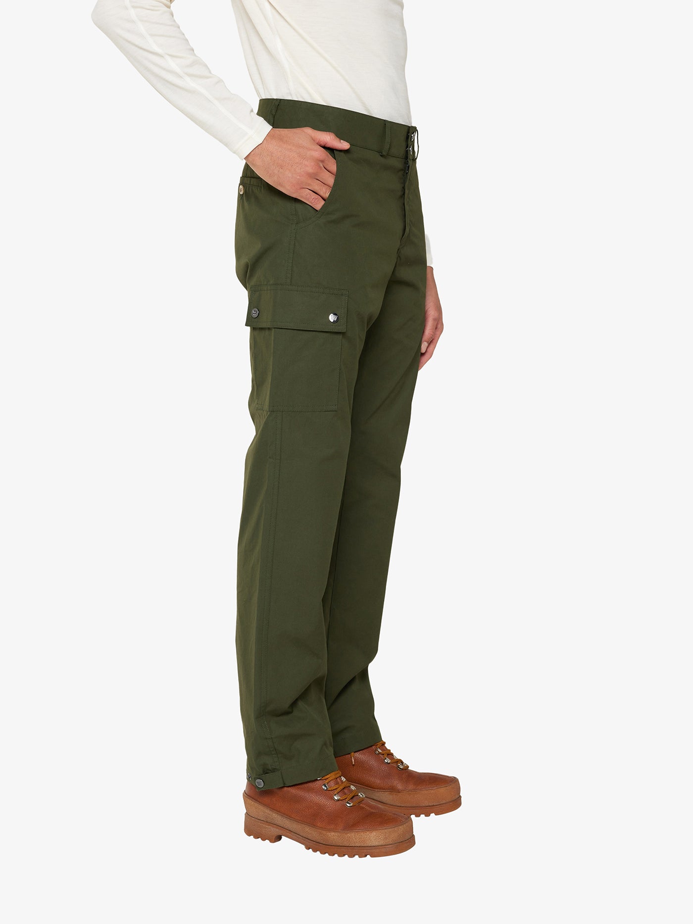 A person wearing We Norwegians' Lyngen Pant Men in olive green, crafted from weatherproof Ventile® cotton fabric with side pockets, paired with brown hiking boots. The person is also dressed in a long-sleeved white shirt. The side view highlights the pants and partially shows one hand in a pocket, making it ideal for outdoor enthusiasts. The background is plain white.