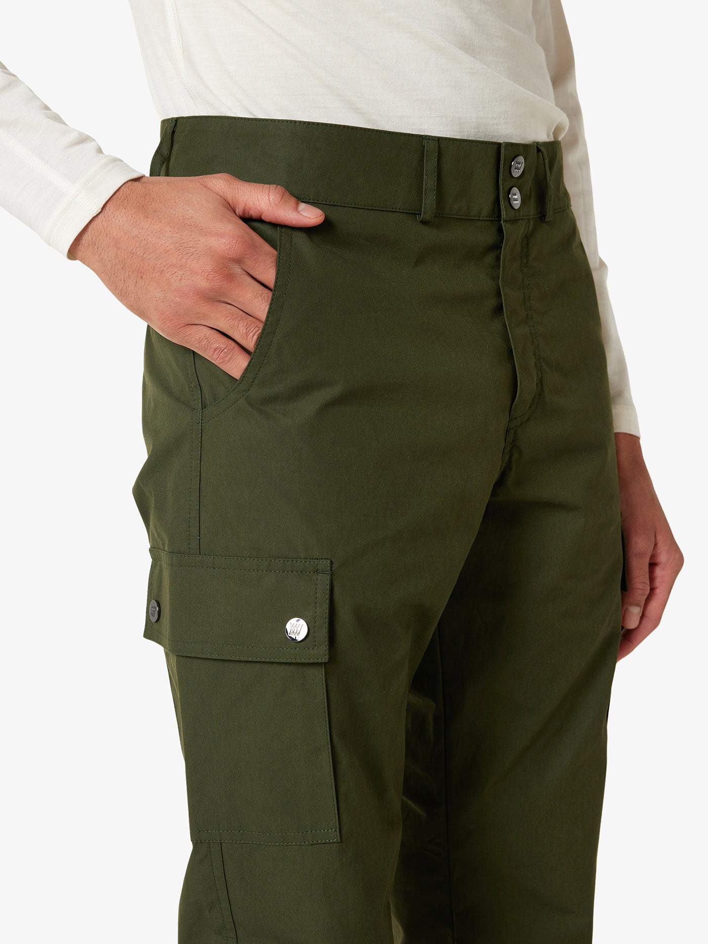 An outdoor enthusiast sports an off-white long-sleeve shirt and the Lyngen Pant Men in Olive Green by We Norwegians, made from durable Ventile® cotton fabric. The weatherproof qualities of these pants, featuring side pockets and buttoned flaps, add both functionality and style. The person has their left hand in one pocket against a plain white background.